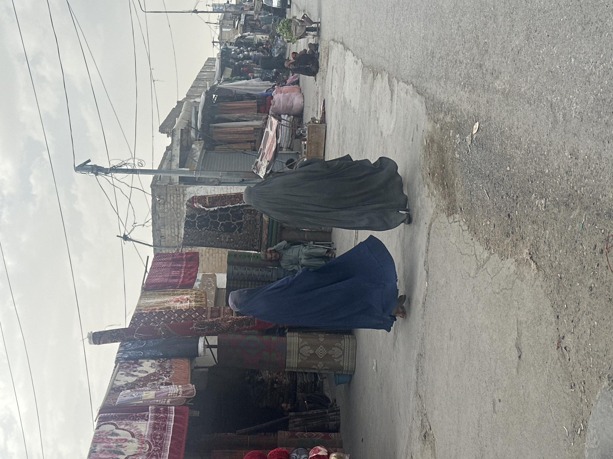 Two women in burqas walk along a concrete sidewalk bordering an outdoor market. The woman in front is wearing a dark blue burqa, and the woman behind her is wearing a dark gray or black burqa. Shops display carpets and textiles. A man sits near the shops on a platform. A crowd of people is visible further down the street. Utility poles with wires are overhead. The scene depicts a street in Quetta City Tehsil, Pakistan.