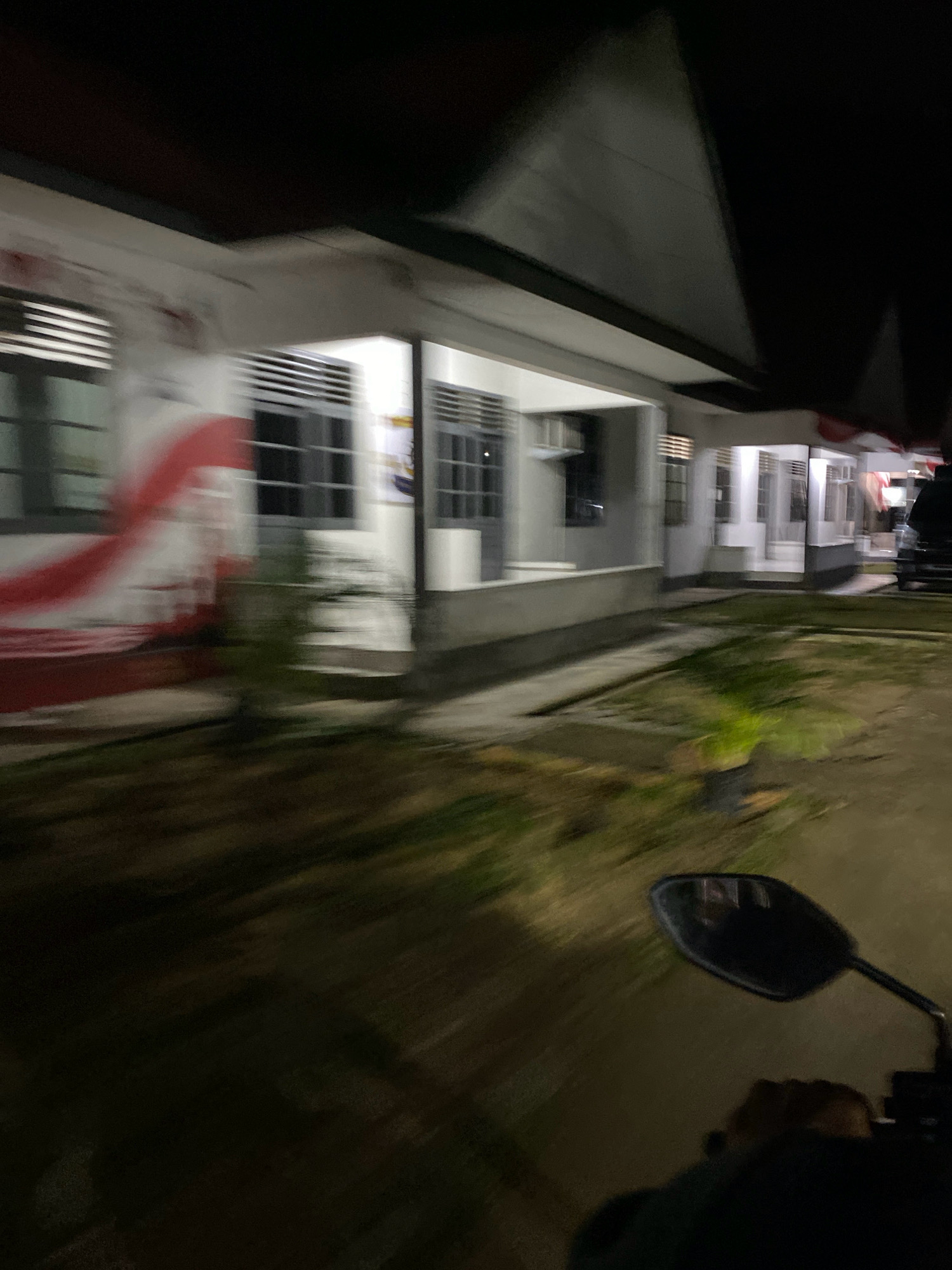 The image depicts a motion-blurred view from the perspective of someone riding a motorcycle at night in Tanjung Pinang, Indonesia. In the foreground, a portion of the motorcycle's handlebar and a rear-view mirror are visible. The background shows a long, low-rise building with multiple windows and a covered walkway. The building's facade is partially obscured by a large red and white mural, resembling the Indonesian flag. Some vegetation, including potted plants, is present on the grounds in front of the building. The scene suggests a street or roadside view in a residential or institutional area of the city.