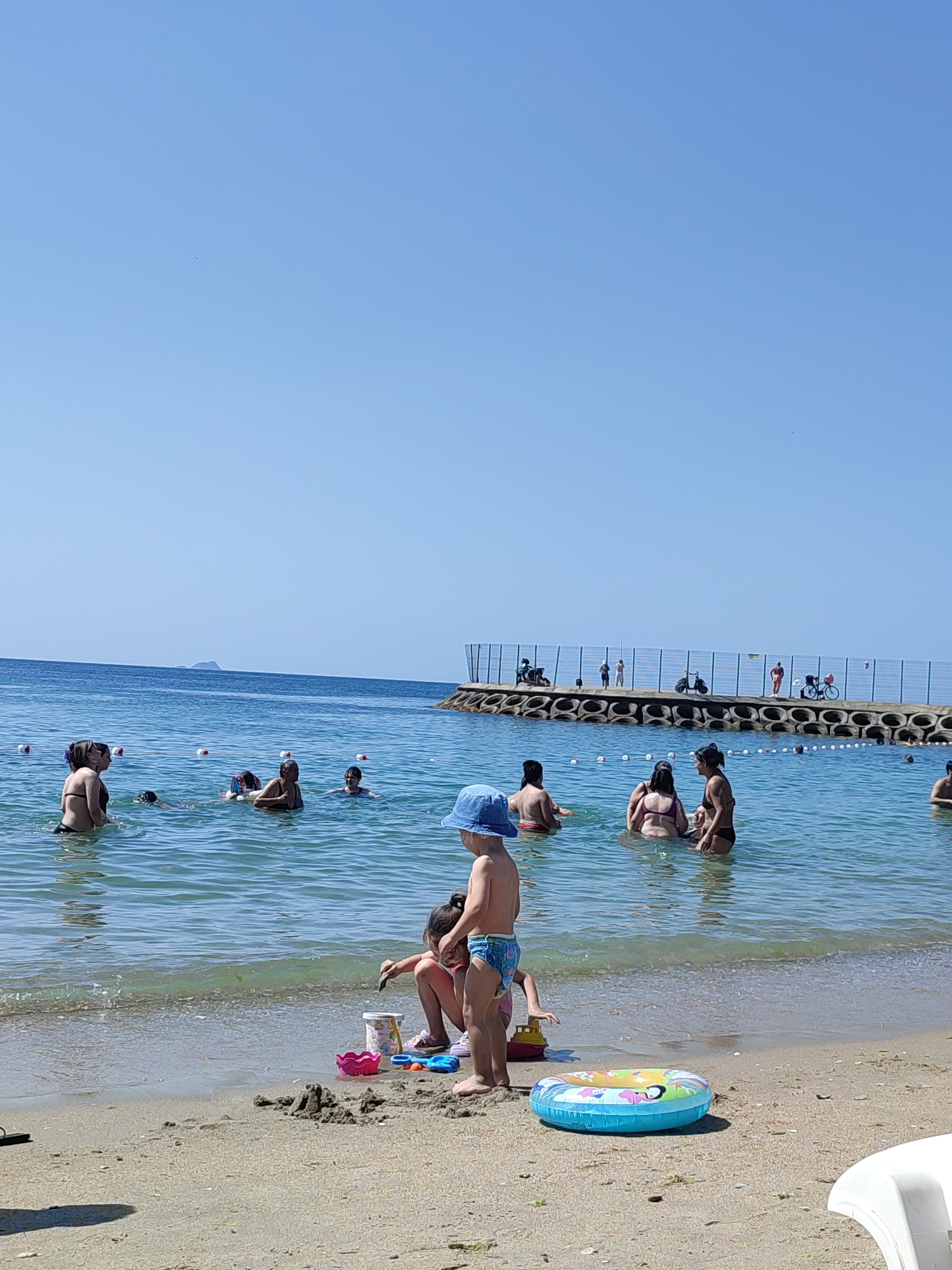 The image depicts a crowded beach scene on a clear, sunny day. The sky is a vibrant blue with no clouds. In the foreground, two children are playing on the sandy beach. One child, wearing a blue sun hat and blue swim trunks, stands facing away from the camera. Another child, partially obscured by the first, is sitting and digging in the sand with a small spade, with a pink shovel and yellow toy boat nearby. A colorful inflatable ring lies on the sand to the right of the children.
The water is calm and clear, with several people swimming and wading. In the middle ground, various individuals are visible in the sea, some standing and conversing, others swimming. Further back, a breakwater extends into the sea, with a few people standing on it. The breakwater is lined with buoys that mark a boundary in the water. The presence of a distant island or landmass on the horizon suggests a coastal location. The overall atmosphere is one of a typical summer day at a popular beach. This scene is consistent with the beaches found along the coast of Kadıköy, Istanbul, Turkey, which are frequented by locals and tourists during the warmer months.