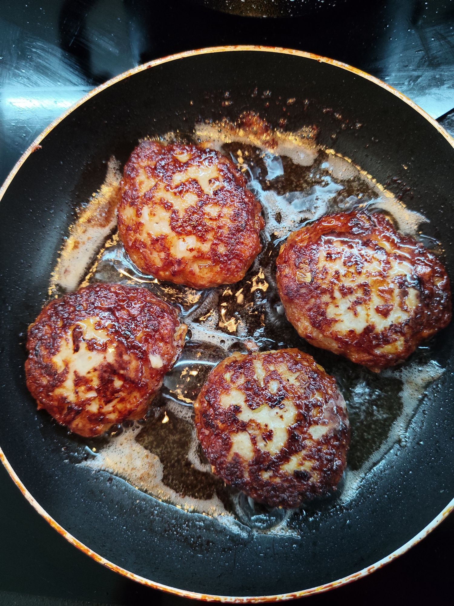 Four hamburger patties are frying in a black skillet on a stovetop. The patties are golden brown and have grill marks. The skillet is filled with oil and butter, which is sizzling and bubbling around the patties. The background of the image is dark and out of focus, suggesting that the focus is entirely on the food. The lighting is bright and harsh, with a strong reflection on the stovetop. The scene appears to be indoors, likely in a kitchen.