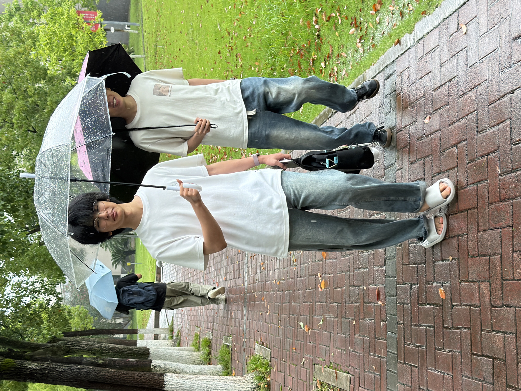 Three individuals are observed walking along a wet, reddish-brown brick pathway in the city of Jiangjin, China, during a rain event. The pathway is laid in a herringbone pattern and shows reflections from puddles, with scattered brown and orange leaves. To the left, a line of trees with white-painted bases runs parallel to the path, displaying green foliage. To the right, a wet grassy area with scattered leaves is visible.
In the foreground, a young male with dark hair and glasses wears a white short-sleeve T-shirt, light blue baggy jeans, and white slide sandals. He holds a clear umbrella with visible raindrops in his right hand and a black shoulder bag with a light blue tag in his left hand. A light pink wristband is on his left wrist. He is looking towards the viewer. Walking to his right, partially obscured, is another young male wearing a light beige short-sleeve T-shirt with a small graphic, blue baggy jeans with a white drawstring, and dark sneakers. He holds a black umbrella with a pink underside in his right hand. Both individuals are sheltering from the rain. In the mid-ground, walking away from the camera, a third individual wears a black T-shirt, khaki cargo pants, and light-colored shoes, carrying a dark blue backpack and holding a light blue umbrella over their head.