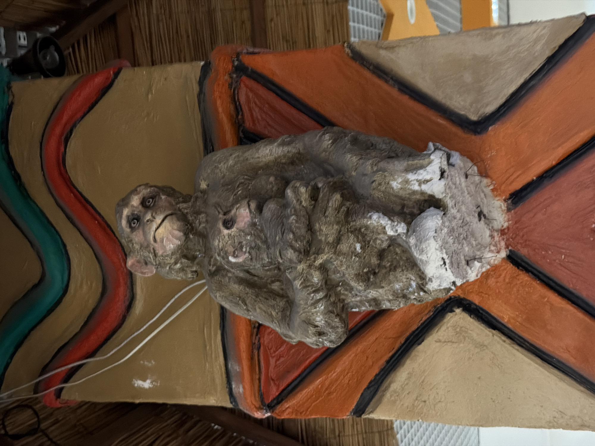 The image shows a statue of a mother monkey holding a baby monkey, affixed to a decorative column. The column is painted with broad, curved bands of teal and red against a tan background, topped with what appears to be thatched roofing. Below the monkey statue, the column is adorned with large, stylized diamond shapes in orange, tan, and red, outlined with black. The statue itself is weathered and appears to be made of a rough material, with the monkeys depicted in a grayish-brown color. The background shows elements of an indoor space, possibly a shopping center or an entertainment venue, with glimpses of white ceiling tiles and orange and yellow accents. The scene is brightly lit. This statue is located at the Kyiv Zoo in Kyiv, Ukraine.