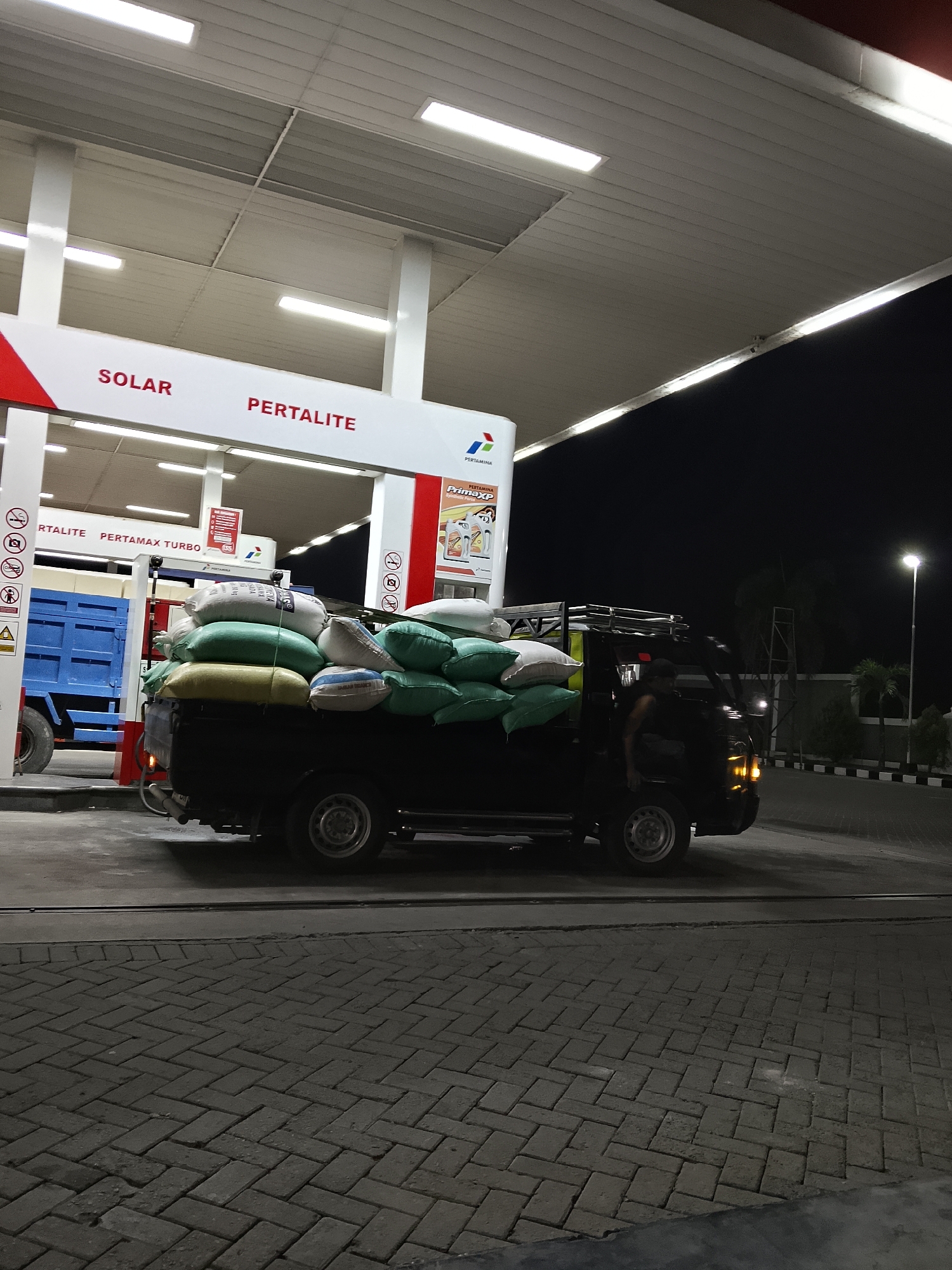The image depicts a nighttime scene at a Pertamina gas station in Kasiman, Indonesia. A black pickup truck is parked under the gas station canopy, loaded with numerous large sacks, predominantly green and white in color. Some of these sacks appear to be filled with grain or agricultural products. A person is visible in the driver's seat of the truck. Adjacent to the pickup truck is a blue truck. The gas station features illuminated signage indicating "SOLAR" and "PERTALITE." The ground is paved with interlocking bricks. A street light illuminates the background. The overall impression is of a commercial transaction or delivery occurring late at night at a roadside fuel stop.