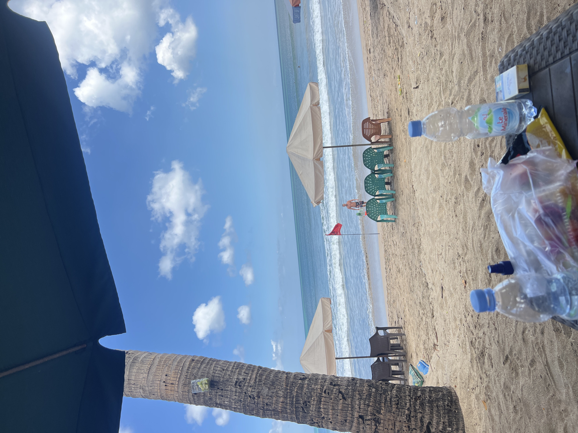 A beachfront scene in Kuta, Indonesia, unfolds under a bright blue sky dotted with white clouds. In the foreground, a dark umbrella casts a shadow, partially obscuring a large palm tree trunk with a clear plastic container holding a few green fruits attached to it. A woven table in the foreground holds a partially filled plastic bottle of "Le Minerale" water, a packet of cigarettes, a plastic bag containing red fruit, and a small dark spray bottle. Beyond the foreground, the sandy beach is occupied by several empty chairs. Two dark brown chairs are positioned under a beige umbrella. Further down the beach, a group of four green plastic chairs and one brown plastic chair are arranged near another beige umbrella. A single red flag stands planted in the sand. In the background, the turquoise ocean stretches out, with gentle waves rolling towards the shore. A man in swimming trunks stands near the water's edge, facing the ocean. The overall scene depicts a relaxed day at a popular tourist destination.