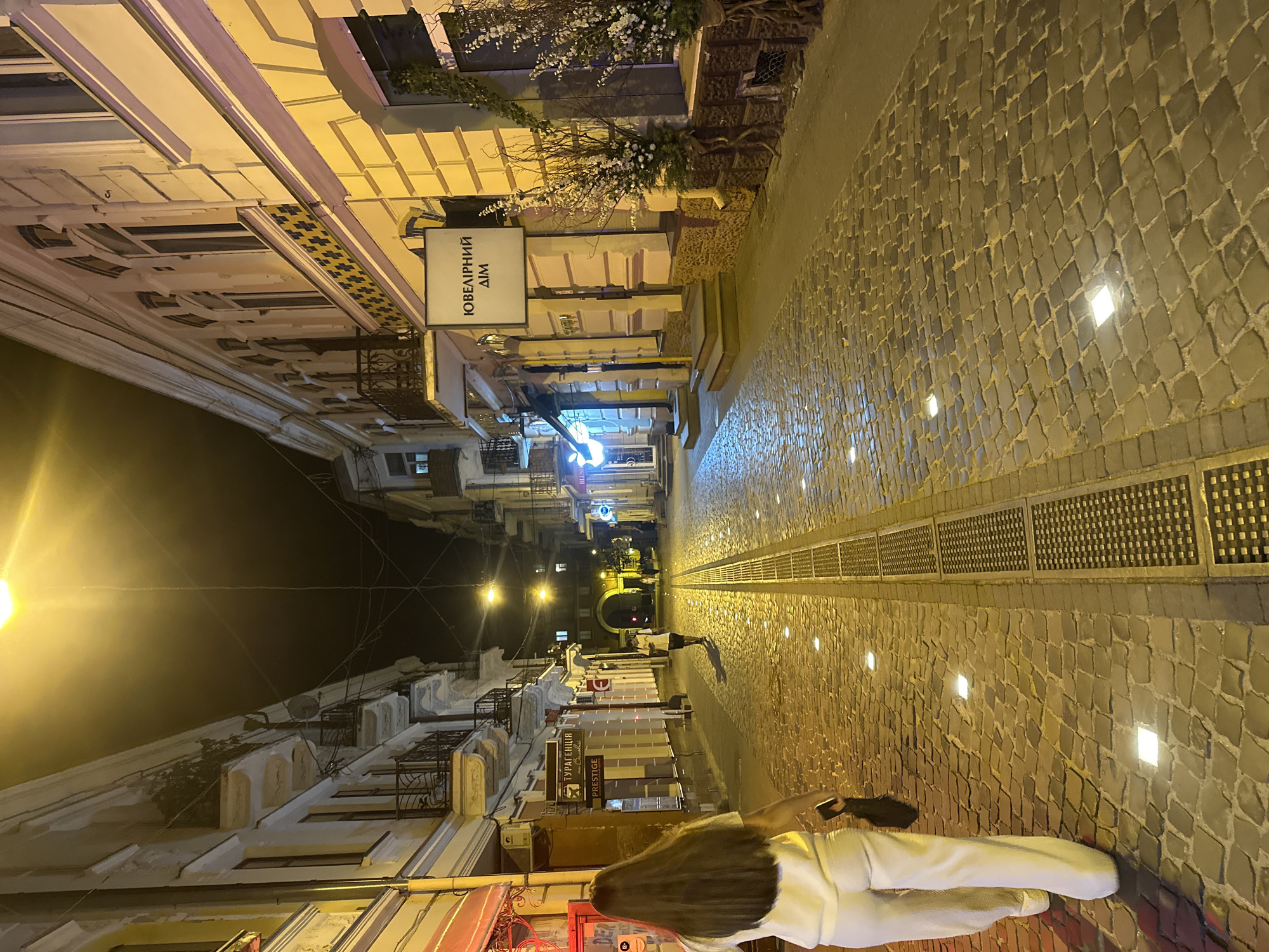 A nighttime street scene in Ternopil, Ukraine, features a cobblestone street illuminated by embedded lights and streetlights. On the right side of the street, a building with a sign reading "ЮВЕЛІРНИЙ ДІМ" (Jewelry House) is visible. On the left side, another building has signs, including one with the text "ПРЕСТИЖ" (Prestige). A woman with long brown hair, wearing white pants and a white top, is walking away from the camera on the left side of the street, carrying a dark bag. Another person is walking further down the street in the distance, and a few more figures are dimly visible in the background. The street is lined with buildings on both sides, and an arched structure is visible at the end of the street.