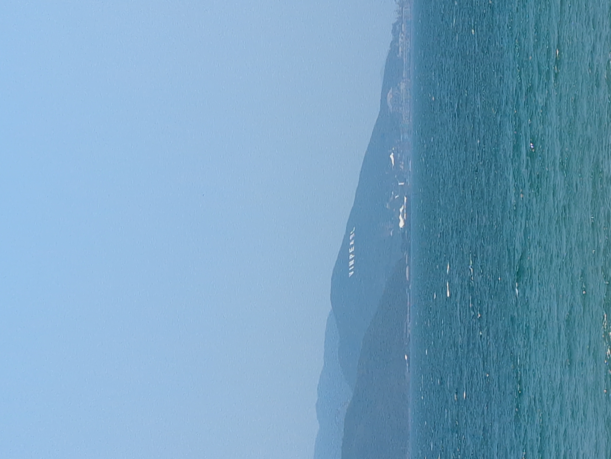 The image shows a scenic view in Nha Trang, Vietnam. The scene consists of a calm, blue ocean on the right side of the image, occupying roughly half of the frame. Adjacent to the ocean, a mountain range stretches upwards, its peaks receding into the distance. On the mountainside, the word "VINPEARL" is inscribed in large, white letters. The sky is clear and light blue, filling the remaining space on the left. The image is oriented in a portrait format.
