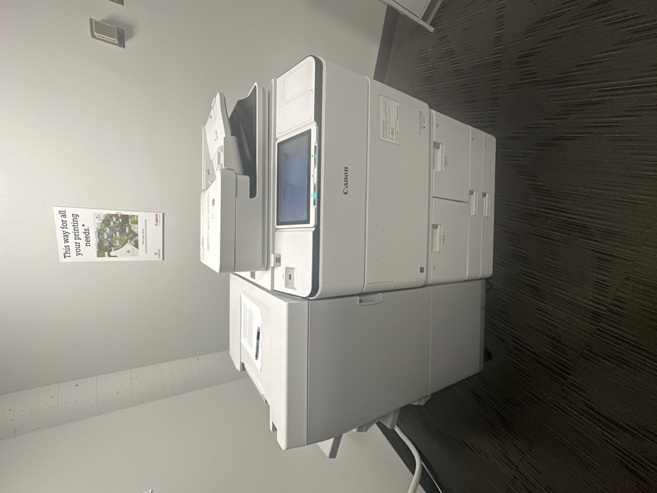 A white Canon imageRUNNER ADVANCE multifunction printer/copier, model P-462, is positioned centrally within an indoor office or service area. The device features a main unit with a central touchscreen display, an automatic document feeder on top, and multiple paper input trays below. A paper finishing unit is attached to its left side, upon which several sheets of paper and a blue pen are placed. The printer rests on a dark gray, subtly textured carpeted floor. The surrounding walls are light gray. A concrete column or pipe is visible in the upper left corner. Directly above the printer, a vertical poster is mounted, displaying the text "This way for all your printing needs." and a graphic depicting a campus map. The poster also includes the text "Clark Center," "Standard for Campus Services Building," and "Canon" and "Epson" brand logos. Two rectangular wall-mounted control panels are visible on the right wall, and a partial view of a white A-frame sign is present on the floor to the right.
The location, indicated by the poster's reference to a "Campus Services Building" and "Clark Center" within the specified city, is likely the Campus Services Building of Wellesley College, Wellesley, United States.