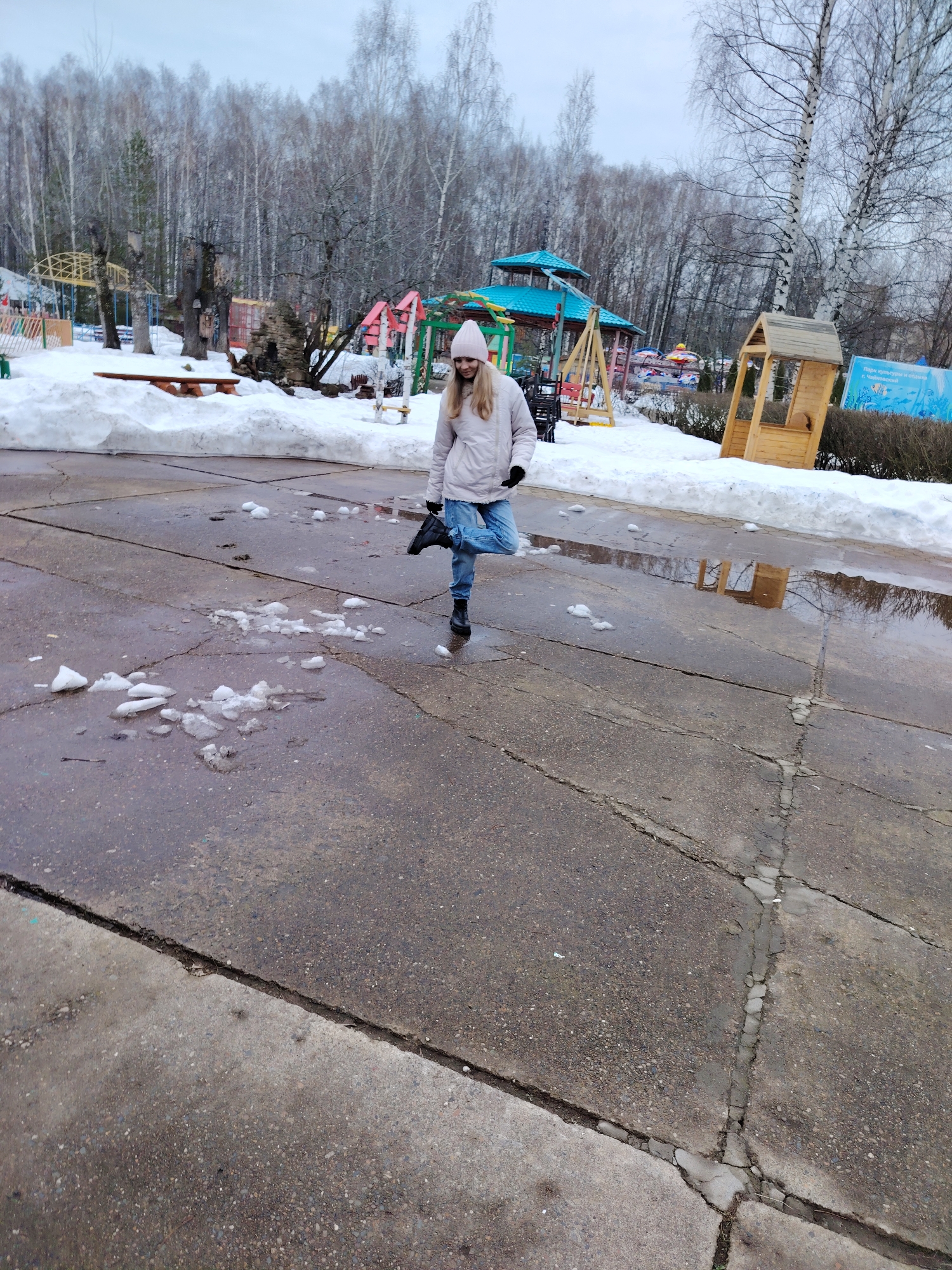 A young woman stands on a wet, cracked concrete surface in what appears to be a park in Chaikovsky, Russia. The woman is wearing a light-colored jacket, blue jeans, and black boots. She also has on a pink hat and black gloves. Her left leg is lifted at an angle toward her body, seemingly kicking at melting ice. The concrete area is interspersed with puddles and patches of melting ice. Behind the woman is a snow-covered area with playground equipment and bare trees. A small wooden structure and a sign with the words "Парк культуры и отдыха г. Чайковский" are visible on the right side of the image. The sky is overcast.