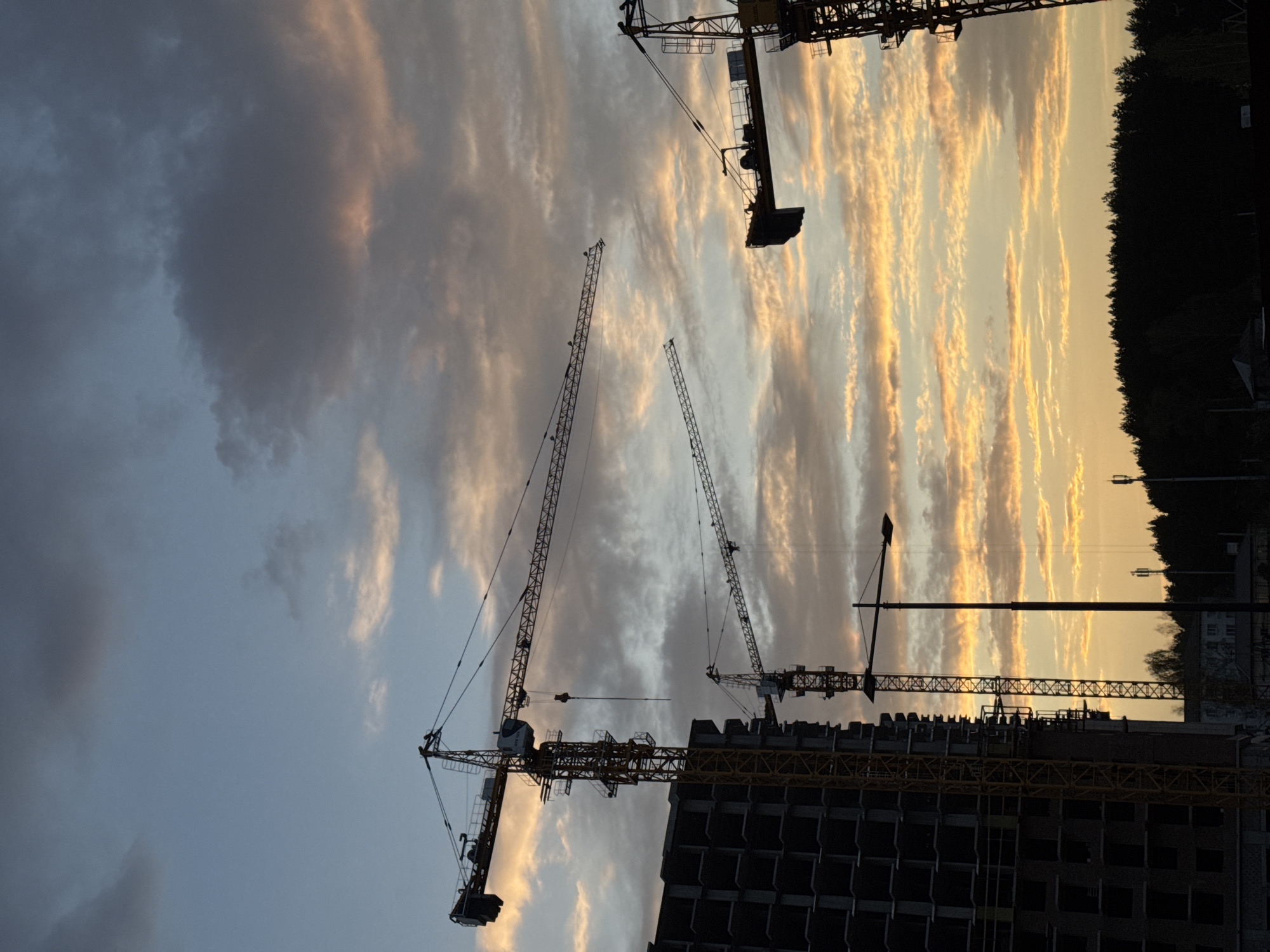 The image shows a construction site with several cranes in the city of Izhevsk, Russia. The cranes are towering over an unfinished building, silhouetted against a dramatic sky with alternating bands of light and dark clouds, illuminated by the setting sun. The environment includes trees in the background.