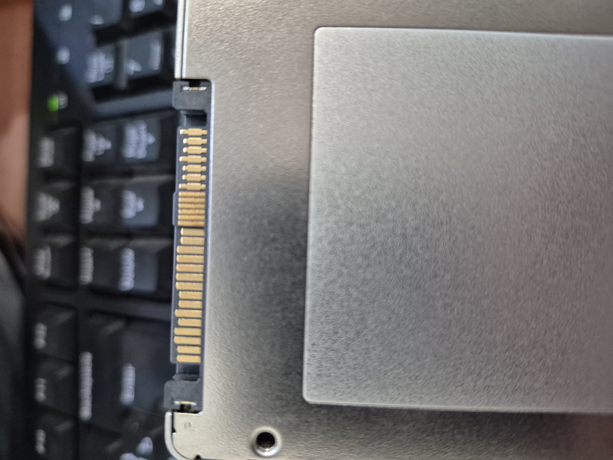 The image shows a close-up view of the connector edge of a solid-state drive (SSD) resting on a keyboard. The SSD has a silver casing and a gold-plated SATA data and power connector. The keyboard is black and appears to be a standard computer keyboard. The background is blurred, but it seems to be an indoor setting. There are no people visible in the image, and no specific details about the city of Homyel, Belarus, can be inferred from the photograph.
