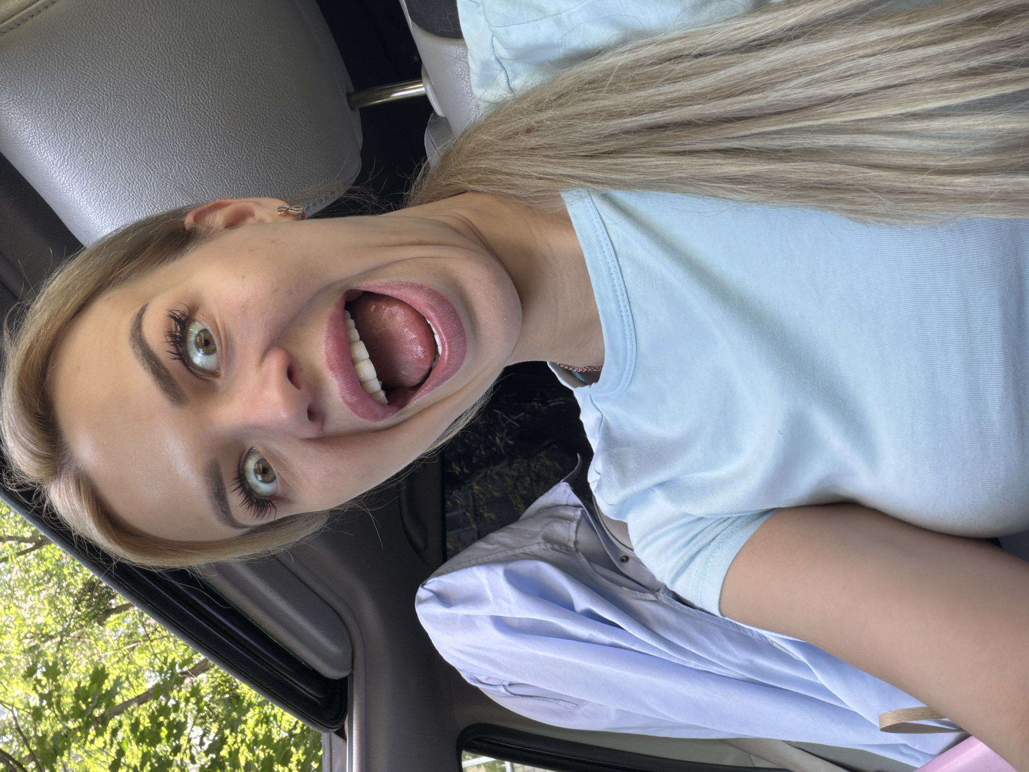A blonde woman with a light blue t-shirt is taking a selfie in a car. She has a surprised expression on her face, with her mouth open wide and her eyes wide. She is wearing a small gold earring in her left ear. A light blue shirt is hanging on the back seat of the car behind her. Sunlight filters in from an open sunroof, illuminating the interior of the car and the trees outside.