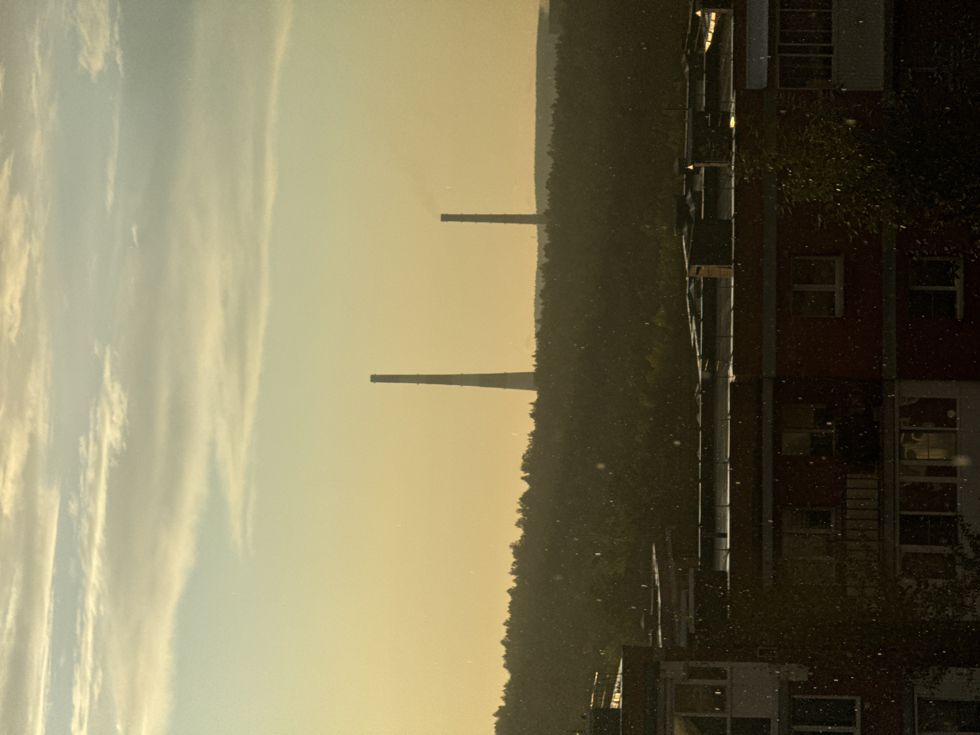 The image depicts a landscape with apartment buildings in the foreground, a densely wooded area in the mid-ground, and two tall industrial chimneys in the background. The sky is a hazy yellow-orange, suggesting either sunrise or sunset, with wispy clouds. There are numerous small white specks scattered throughout the image, which could be dust, pollen, or precipitation. The apartment buildings are multi-story structures with visible windows and balconies. The wooded area appears to be a dense forest. The industrial chimneys are tall and slender, with the one on the left being taller and more prominently visible. The overall scene suggests an industrial or semi-industrial area on the outskirts of a city, with the city of Маркова, Russia, being the specified location.