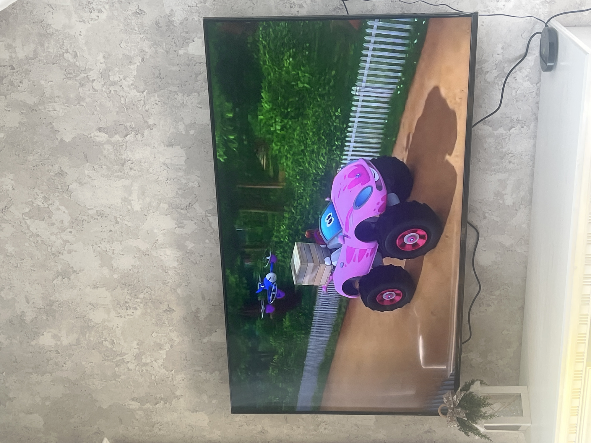 The image shows a television displaying a cartoon. The cartoon features a pink monster truck-like vehicle with a blue character in the cab, positioned next to a small drone. The background of the cartoon includes a green grassy area and a white picket fence. The television is mounted on a textured gray wall. To the right of the television is a white cabinet. There is a small decoration on the cabinet that is decorated with pine needles. The image was most likely taken inside a home, but there is no evidence in the image to indicate this image was taken in the city of Yerevan, Armenia.