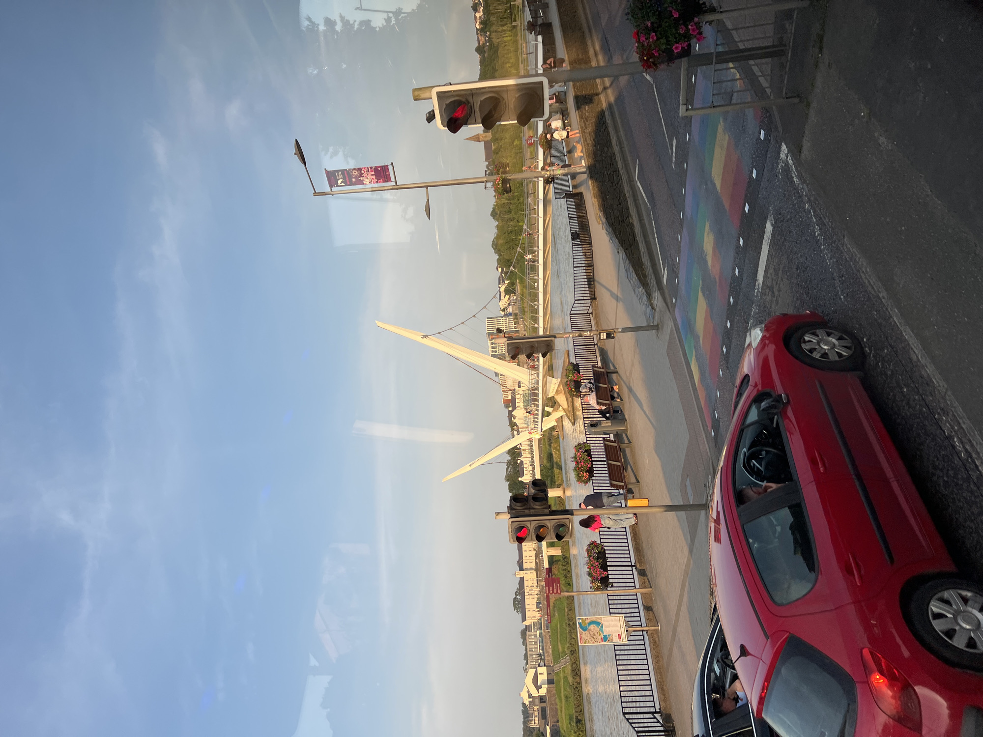 This media file shows an outdoor scene with a river and a pedestrian bridge. The image is captured from a slightly elevated perspective, possibly from a vehicle or a window, and it's oriented vertically.
In the foreground, a red car is parked on the right side of the image, facing away from the viewer. The car's driver-side window is open, and a person is visible inside. Next to the car, a section of road displays a rainbow-colored pedestrian crossing.
The middle ground features a riverbank with people walking and sitting on benches. A distinctive white, modern pedestrian bridge stretches across the river in the background. Traffic lights are visible along the riverbank, with at least one showing a red light.
The background shows buildings on the other side of the river and a treeline. The sky is blue with some wispy clouds, suggesting it is daytime, likely late afternoon or early evening due to the warm lighting. There is a flag with text and a logo hanging from a lamppost near the traffic lights. Another sign with a map is also visible. The scene appears to be in an urban or semi-urban environment, with the rainbow crossing possibly indicating a location known for its inclusivity or a specific event. The overall mood is calm and ordinary, depicting a typical day by the river.