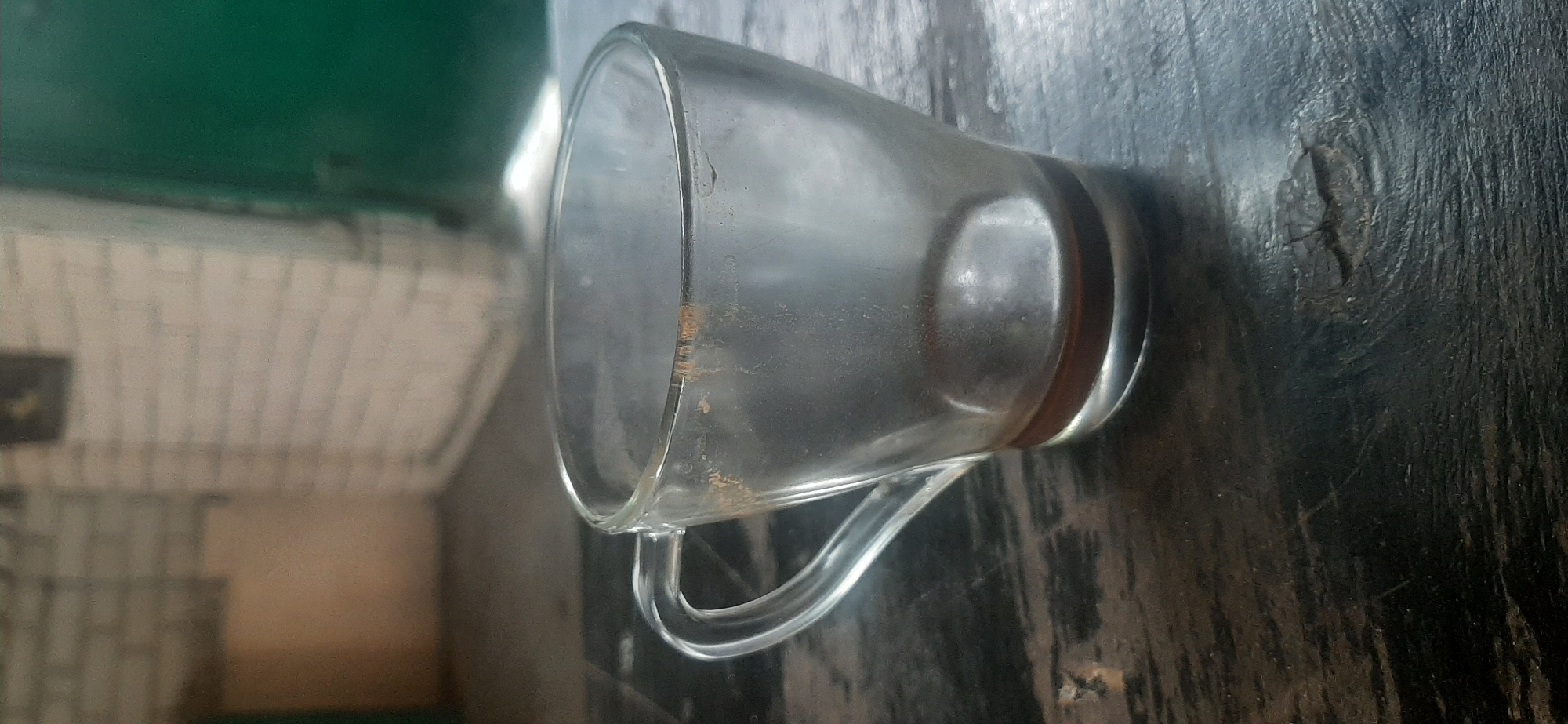 This media file features a clear glass mug with a handle, positioned on a dark, textured surface that appears to be wood. The mug is empty and slightly dusty, with some faint brown residue visible on the inside rim. There is a visible reddish-brown band around the exterior base of the mug. The background is blurred, showing a green surface above a striped white and beige pattern. The lighting suggests it's daytime. The overall scene is a still life, with the focus on the mug and the surface it rests on. There are no people or any discernible activity taking place. The texture of the dark surface is prominent, showing grain and some imperfections, possibly indicating an outdoor table or a rustic indoor setting in Yessentukskaya, Russia. No text is visible in the image.