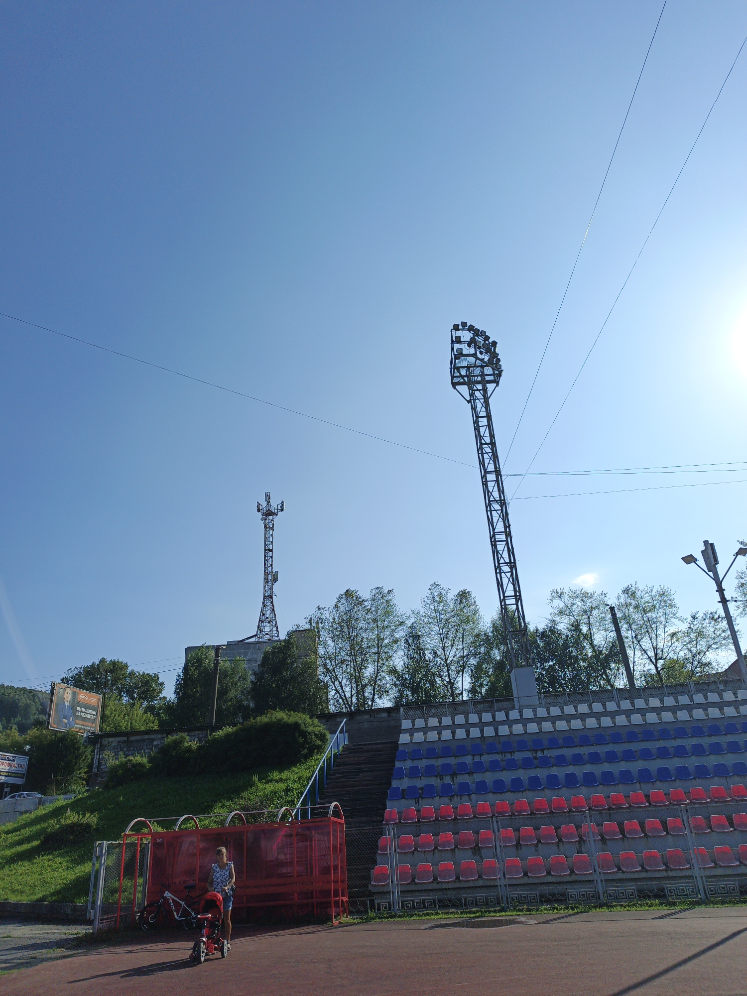 The image depicts a scene at a sports stadium, likely the Metallurg Stadium in Zlatoust, Russia, given the presence of stadium seating and floodlights. A young girl is standing near a red metal fence, holding the handle of a tricycle, with a bicycle leaning against the fence. Behind her, rows of stadium seats are visible, arranged in alternating rows of red, blue, and white. Two tall towers are visible in the background: one is a stadium floodlight, and the other is a communication tower with antennas. The sky is clear and blue, with the sun brightly shining on the right side of the image, causing some lens flare. The environment appears to be a sunny day, with green foliage and trees surrounding the stadium. A billboard advertising a political figure is visible on a hillside in the upper left portion of the image.