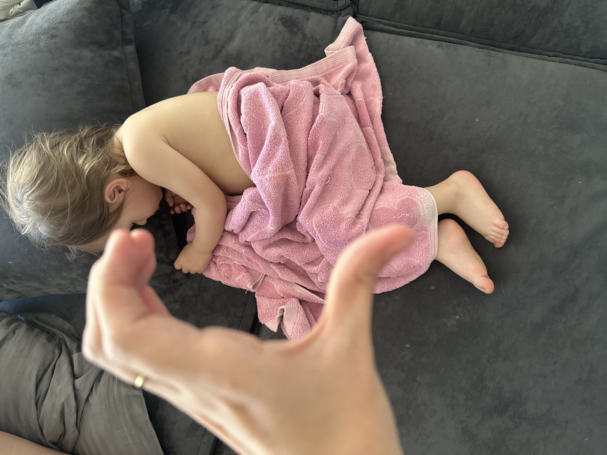 A child, possibly a toddler, is lying on a dark grey sofa, mostly covered by a pink towel. The child is lying on their side with their head near the left edge of the sofa. The child's feet are visible near the right edge of the sofa. A hand is raised in the foreground, partially obscuring the view. The location and city of Kharnavy, Russia, are not indicated in the image.