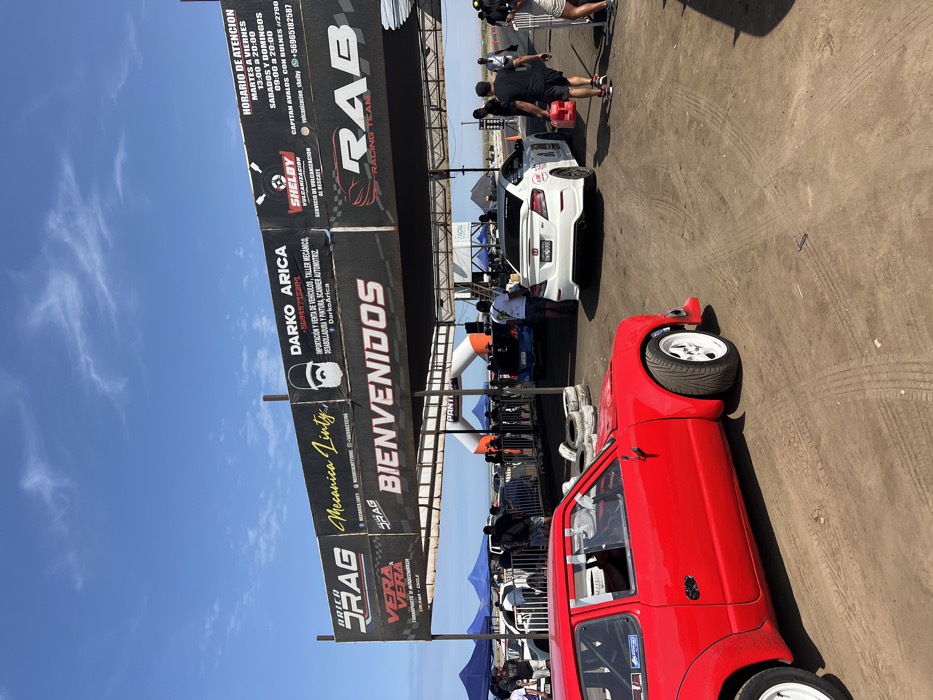 A sunny day at a drag racing event features two cars: a red hatchback in the foreground and a white Honda Civic further back. Sponsors' banners arch over the track, advertising "ARICA DRAG", "RAB RACING TEAM", "DARKO ARICA", and "SHELBY VULCANIZACION". Spectators and crew members are visible near the white car. The track surface shows tire marks, and the sky is clear with a few wispy clouds.