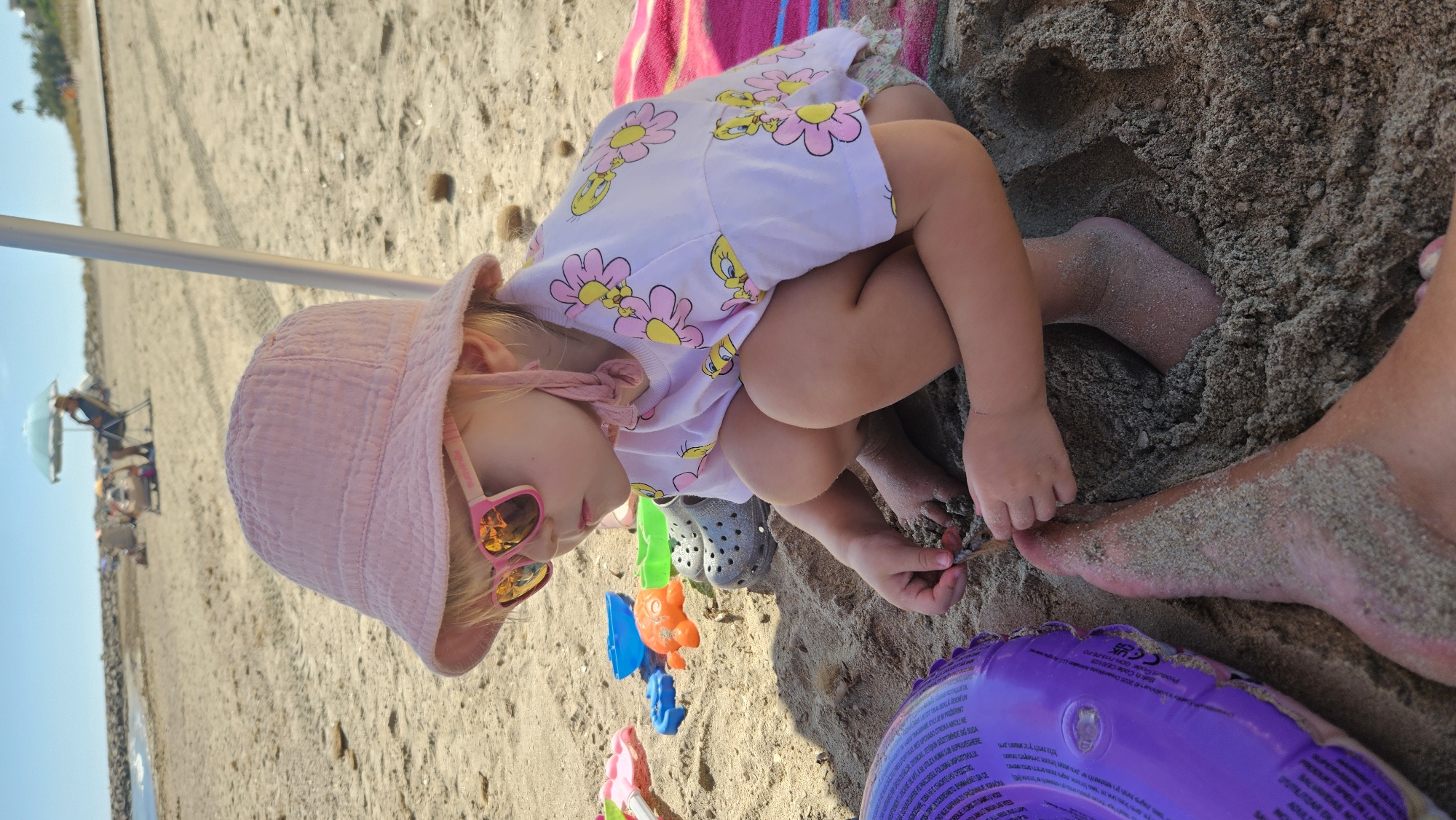In Deveses, Spain, on a sandy beach under a bright sky, a young child with blonde hair is crouched down, engaged in playing with sand. The child wears a pink bucket hat, pink sunglasses with an orange tint, and a light pink t-shirt adorned with a pattern of yellow birds and pink flowers. Nearby, several colorful plastic beach toys are scattered in the sand, including an orange crab-shaped toy and a blue bucket. A gray Croc shoe is also visible. In the foreground, the leg and foot of an adult are partially submerged in the sand, next to a purple inflatable ring. In the background, other beachgoers are present, some sitting under a beach umbrella. The overall scene depicts a relaxed day at the beach.