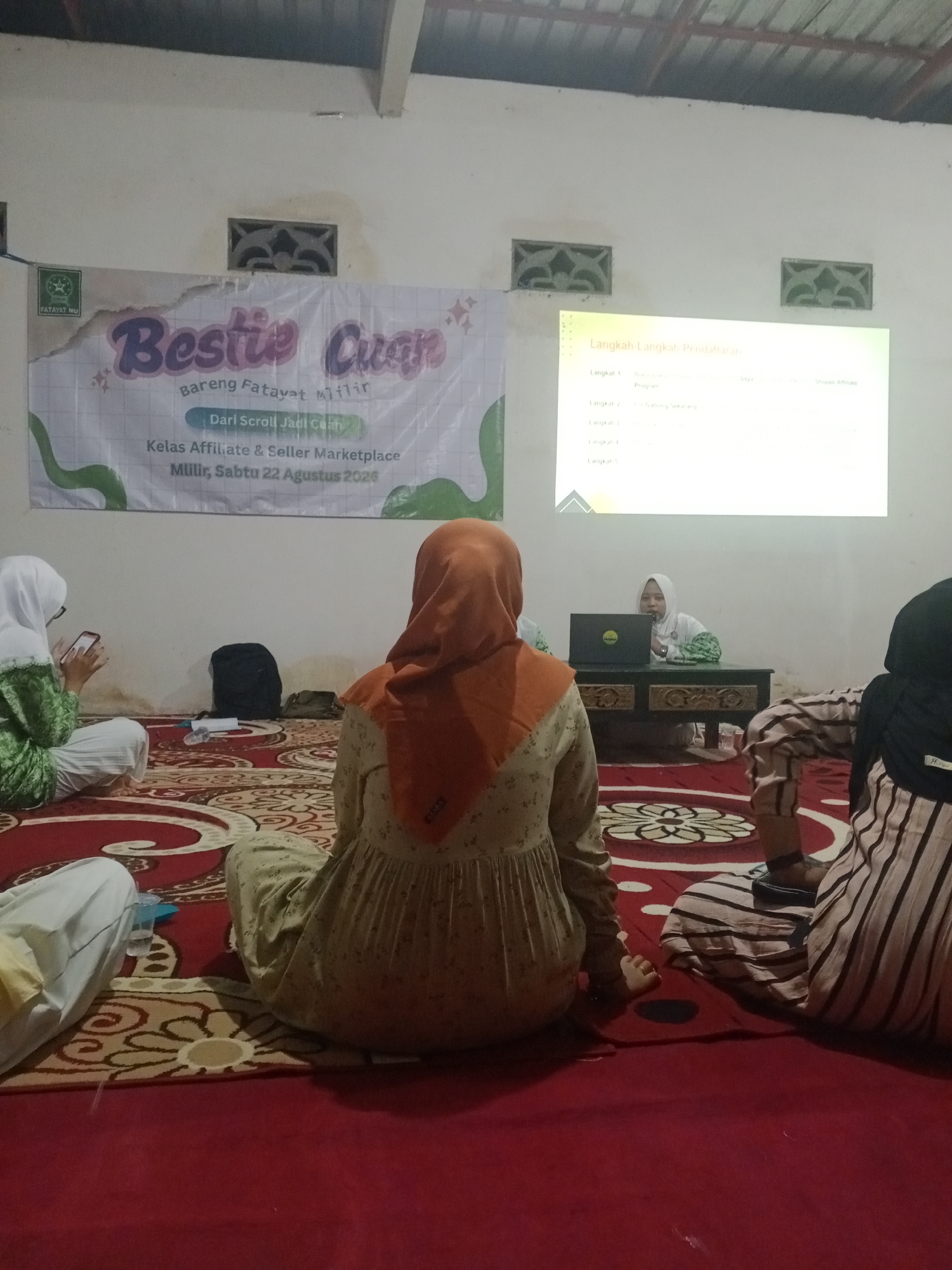 In an indoor setting, likely a community hall or classroom in Mlilir, Indonesia, a group of women are gathered on the floor, seated on a patterned rug. They are attending a class titled "Bestie Cuán: Bareng Fatayat Milir - Dari Scroll Jadi Cuan. Kelas Affiliate & Seller Marketplace, Milir, Sabtu 22 Agustus 2024". The banner announcing the event is visible on the wall behind them.
A projector displays a presentation titled "Langkah-Langkah Pendaftaran" (Registration Steps) on a screen, showing a list of numbered steps with associated actions and platforms. A woman, dressed in a white hijab and top, is seated at a table in front of the screen, appearing to present or facilitate the session, with a laptop open before her.
Several women are visible from behind, one in a light green and white patterned outfit and hijab, looking at her phone, and another in an orange hijab and a light-colored patterned dress. Another individual, partially visible, wears a black hijab and a striped outfit. The environment appears informal, with a low ceiling and ventilation grates on the wall. Bags and a water bottle are also present on the floor. The overall scene depicts an educational gathering focused on online business and marketplace strategies.