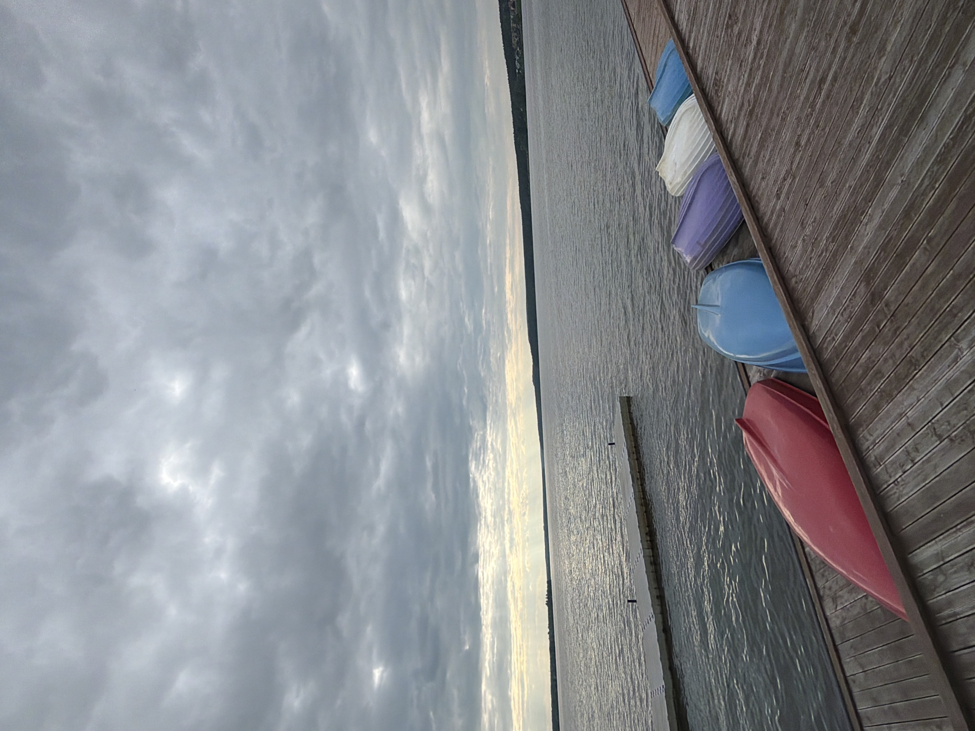 The image depicts a serene lakeside scene. In the foreground, a wooden dock extends into the calm water, with several colorful kayaks and rowboats resting upside down on its surface. These boats are red, blue, purple, white, and light blue. Beyond the dock, a long, flat platform juts out into the lake. The lake itself is wide and reflects the overcast sky, which is filled with dramatic, textured clouds. In the distance, a tree-lined shore forms the horizon, with faint signs of human habitation visible. The overall atmosphere is tranquil, suggestive of a quiet day by the water. This scene is located on the shore of Lake Votkinsk in the city of Votkinsk, Russia. There are no people visible in the image.
