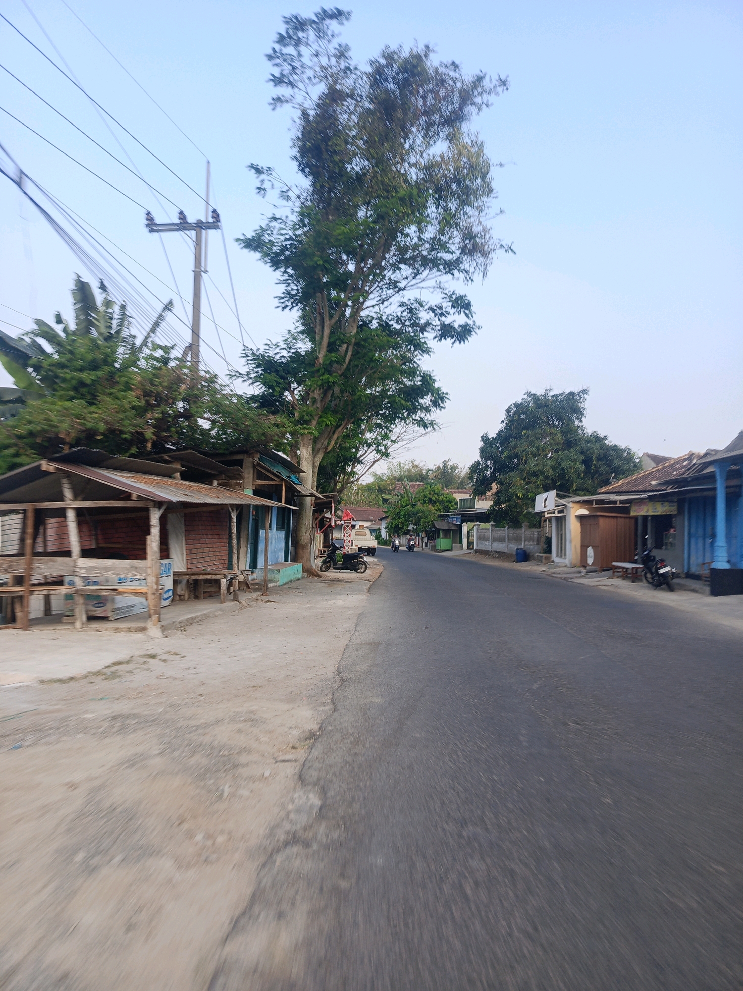 The image depicts a roadside scene in the city of Sumberagung, Indonesia. The road, paved with asphalt, extends into the distance. On the left side of the road, there are several small, rustic structures, appearing to be market stalls or small shops. These structures have tin roofs and are made of wood and brick. Some are open-fronted, displaying wooden planks and possibly building materials. Behind these structures, a large tree with dense green foliage stands prominently. To the right of the road, more buildings are visible, some with blue-painted facades and tiled roofs. A black motorcycle is parked in front of one of these buildings. In the background, other trees and buildings suggest a semi-rural or suburban environment. Overhead, power lines supported by utility poles stretch across the frame. The sky is clear and pale blue, indicating daytime. A few people are visible further down the road, some on motorcycles, suggesting normal activity in the area. The overall impression is one of a typical, somewhat weathered streetscape in a warm climate.