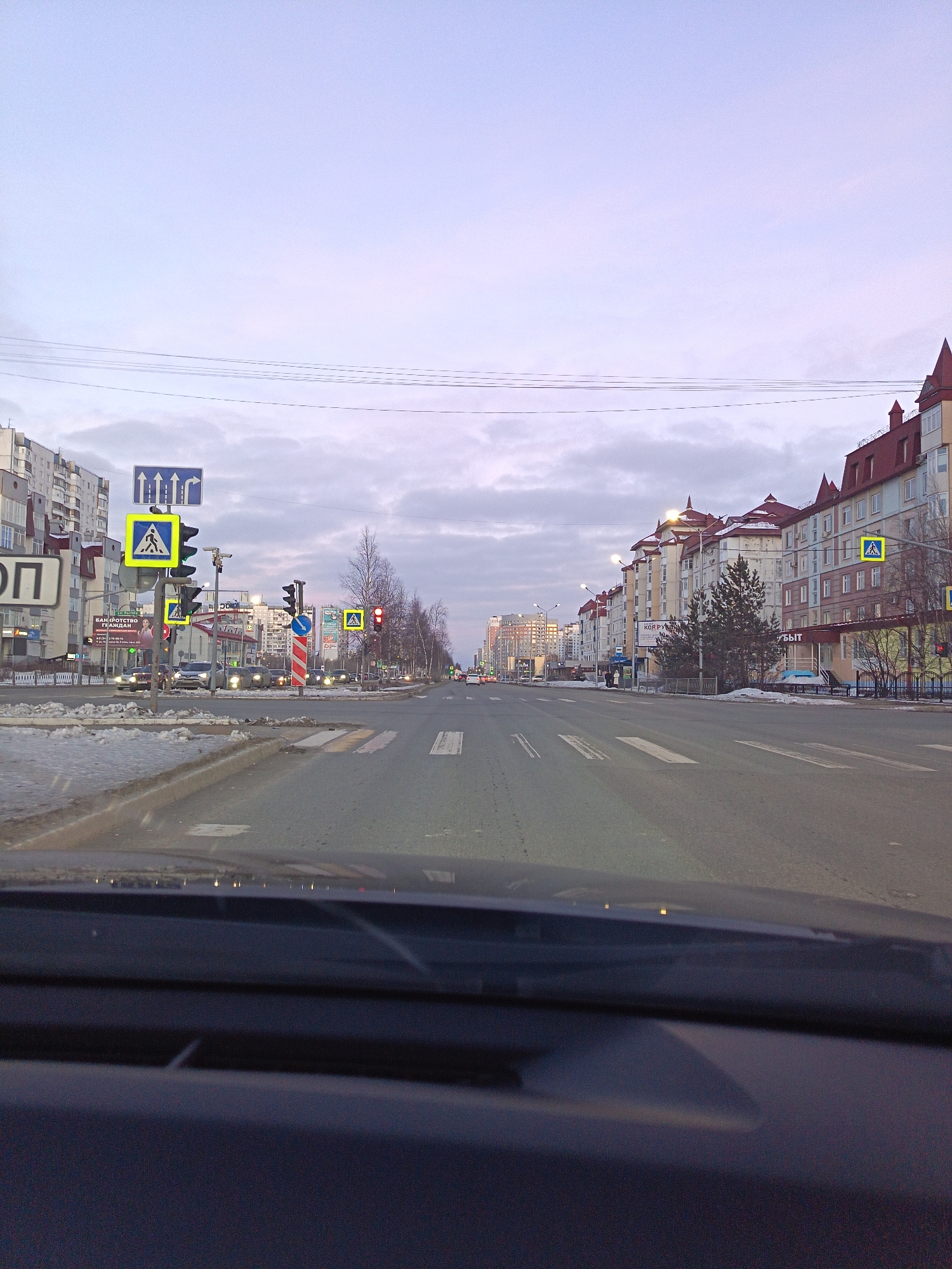 The image shows a view from inside a vehicle driving along a street in Nizhnevartovsk, Russia. It is daytime with overcast skies. There are buildings on both sides of the road, some are high-rise apartment complexes. Pedestrian crosswalk signs are visible. Cars are present in the distance at a traffic light. There are remnants of snow along the side of the road and on the sidewalks. Traffic signals are visible, some showing red lights.