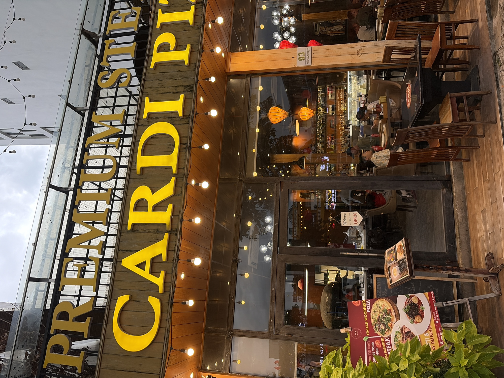This is a street-level, outdoor, eye-level medium shot of the exterior of "PREMIUM STEAK CARDI PIZZA" restaurant. The restaurant has a prominent yellow neon sign with large block letters spelling out the name of the establishment. The sign is mounted above the entrance, which consists of large glass doors and windows, allowing a view into the brightly lit interior. Strings of decorative lightbulbs hang across the storefront.
To the left of the entrance, a stand displays a large, colorful advertisement for "ITALIAN WOODFIRED PIZZA" and "PREMIUM IMPORTED STEAK," with images of food and text detailing the offerings. Next to the advertisement stand is a wooden menu holder displaying a small menu.
Inside the restaurant, patrons are seated at tables, some of which have pizzas. The interior appears to be well-lit, with shelves of bottles visible behind the counter area. A sign near the entrance reads "Welcome" and "Open." A small plaque on the right side of the entrance has the number "93" and text that appears to be a Vietnamese address, likely "Võ Văn Kiệt," indicating the location is in Đà Nẵng, Vietnam. The scene suggests a lively evening at a restaurant.