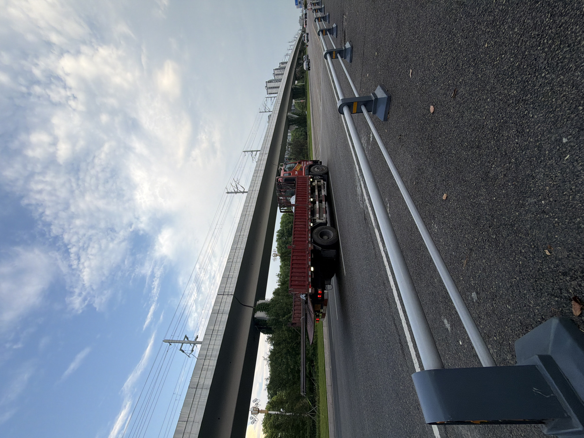 The image captures a scene on a multi-lane road under an elevated railway line. A red flatbed truck is moving in the right lane, carrying long, dark cylindrical objects on its bed. The truck has a metal railing along its sides. The elevated structure, supported by concrete pillars, features overhead electrical wires for the train. In the background, more urban infrastructure and buildings are visible, along with a few other vehicles on the road. The sky is partly cloudy with a mix of white clouds and blue sky. A silver metal guardrail with gray posts runs along the right side of the road, in the foreground. The road surface is asphalt. The setting appears to be a suburban or semi-urban area, likely part of the city of Jiangjin, China, given the architectural style and the presence of elevated rail infrastructure commonly found in Chinese cities. There are no people clearly visible in the image.