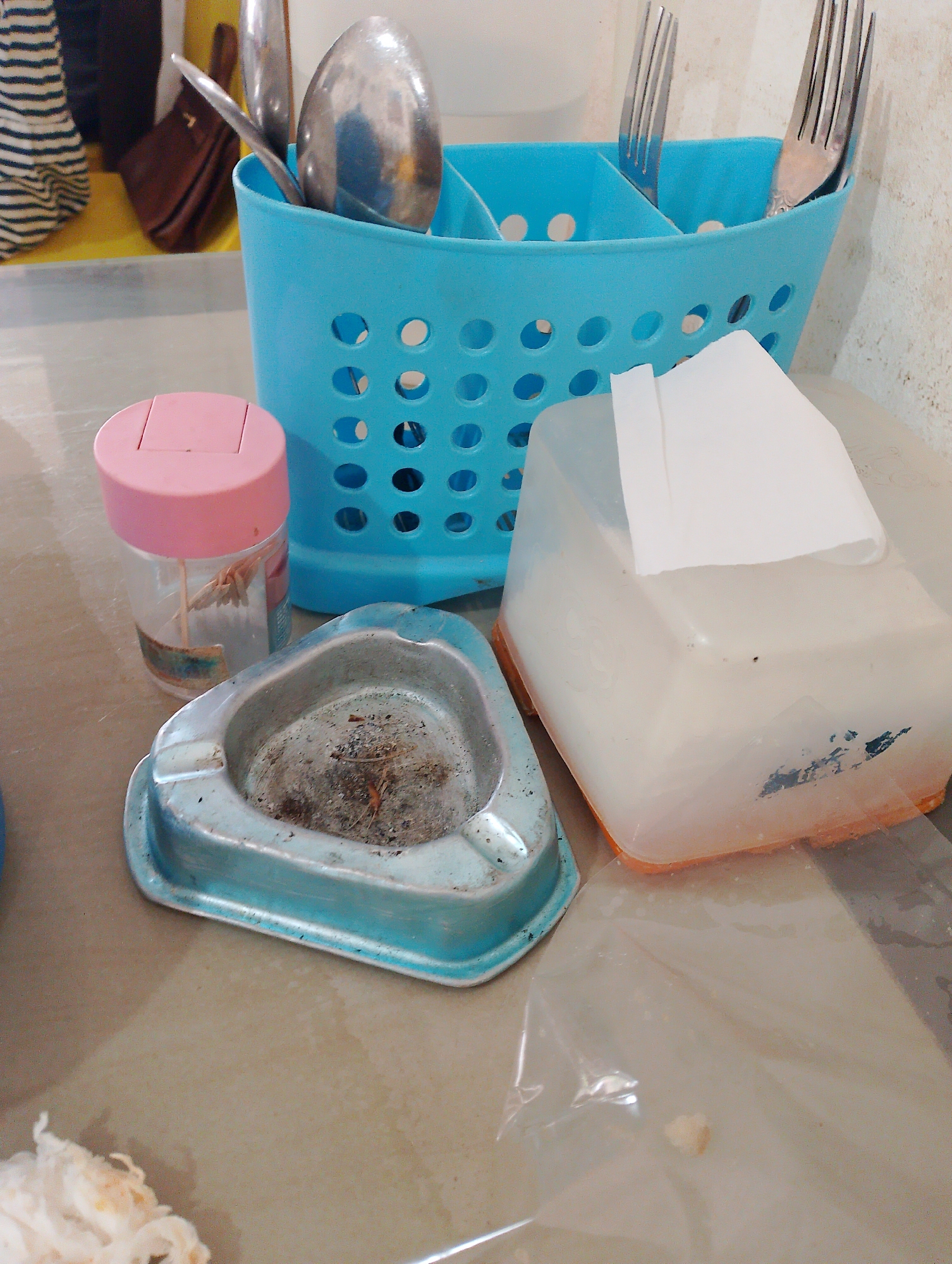 A close-up, eye-level shot displays a collection of items on a neutral-colored surface, possibly a countertop in Batam, Indonesia, given the context. Dominating the center is a bright blue plastic utensil holder filled with metallic cutlery, including spoons and forks. To the left, a small, clear plastic container with a pink lid holds what appears to be toothpicks, with a single toothpick visible inside. Adjacent to this is a light blue, triangular ashtray containing ash and debris, suggesting recent use. In the foreground and right of the ashtray, a translucent plastic tissue box with an orange base is partially visible, with a white tissue pulled out. The background features a blurred image of what could be a yellow tray and a brown handbag. The lighting is soft, and the overall scene suggests a casual, perhaps domestic or roadside eatery setting in Batam.