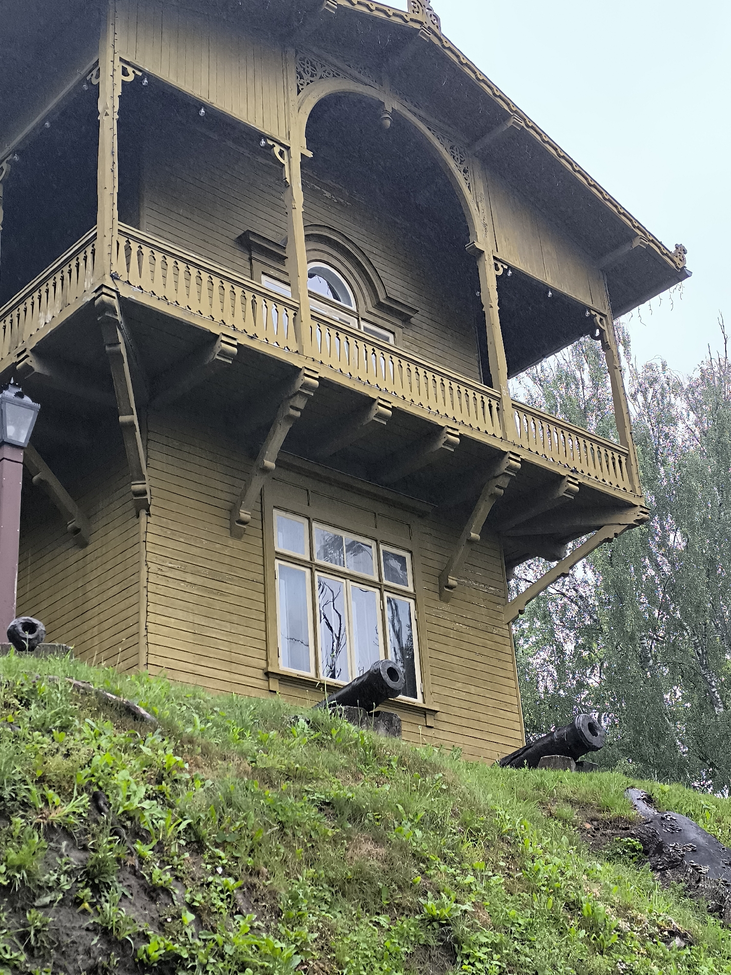 The image shows a two-story wooden building with a distinctive balcony, painted in a muted olive green. The building has a prominent balcony on the upper level, supported by decorative brackets and featuring a wooden balustrade. An arched window is visible on the upper floor, and a large, multi-paned window is on the lower floor. The building is situated on a grassy slope. In the foreground, on the slope, are two old cannons. The overall scene suggests an historical site. The building and cannons are located in Kuldīga, Latvia, specifically at the Kuldīga Castle Mound, a historical defensive structure. The sky is overcast, and there appears to be some precipitation, possibly rain or snow, visible as small white specks in the air. The surrounding trees are lush green, indicating a season of growth.