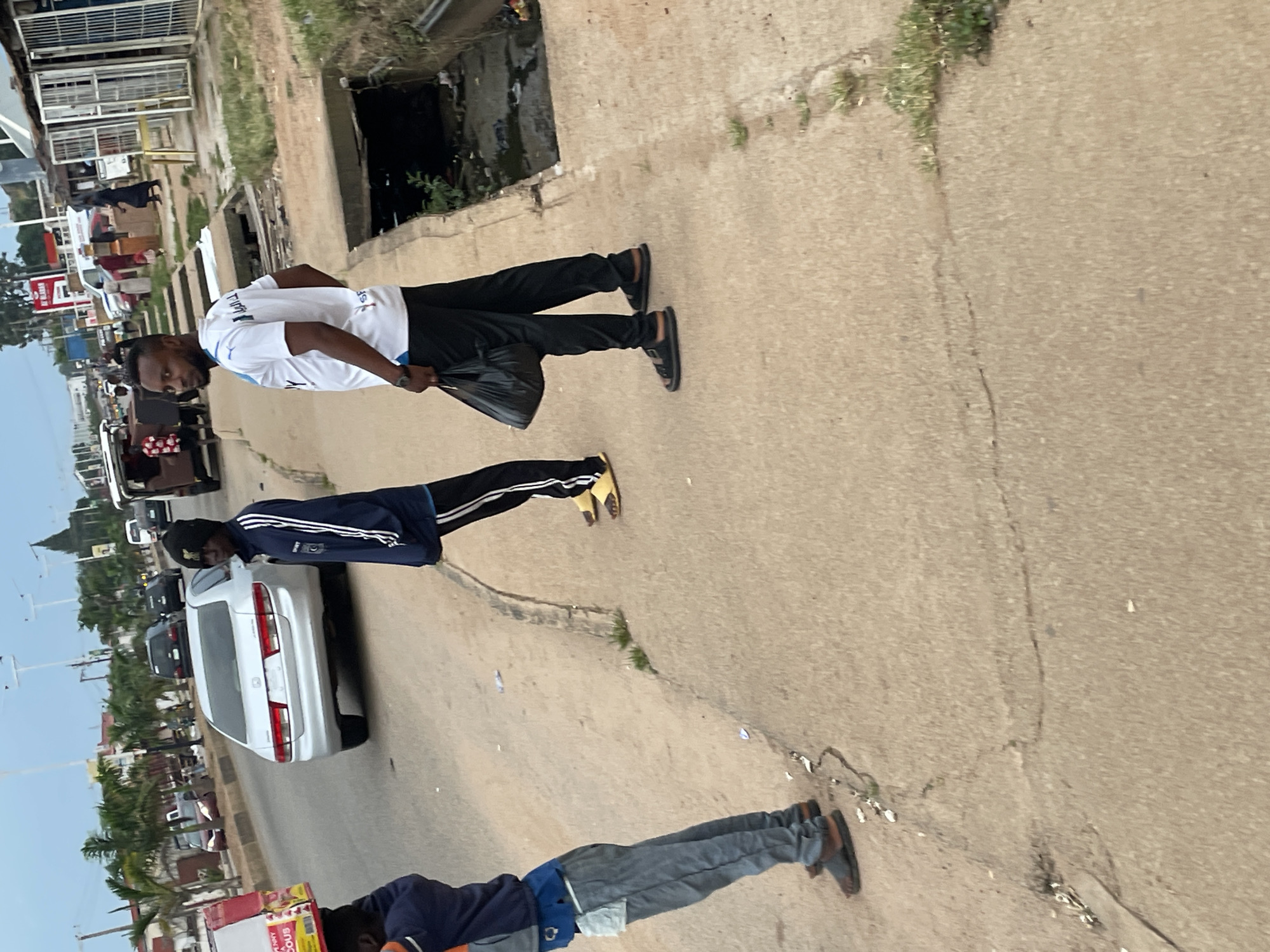 In Barakin Lerawi, Nigeria, a group of individuals stand on a paved walkway adjacent to a road. In the foreground, a person wearing a blue jacket and grey pants carries a large cardboard box on their head. To their right, another person in a blue jacket and black pants with yellow stripes walks away from the camera. Further to the right, a man in a white t-shirt with blue trim and black pants holds a black plastic bag. Behind them, a silver sedan is parked on the road. The environment includes a grassy verge with a drainage ditch, and various buildings and palm trees are visible in the background, suggesting a commercial or residential area. The sky is overcast.