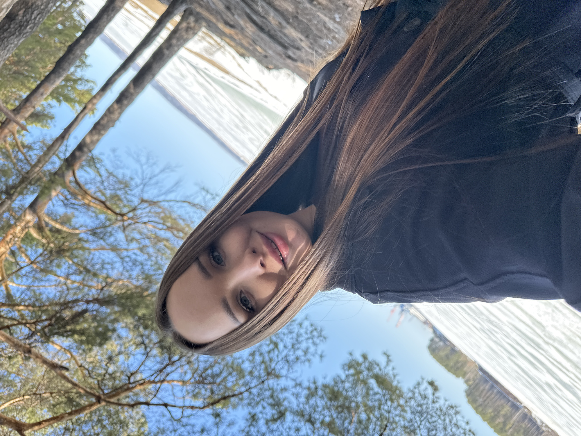 A young woman with long brown hair is in the city of Chaikovsky, Russia. She is facing the camera, with bare trees and blue sky visible in the background. In the top right corner, there is a body of water or ice with some trees reflecting in it. The image is oriented so that the woman's head is slightly tilted to the right.