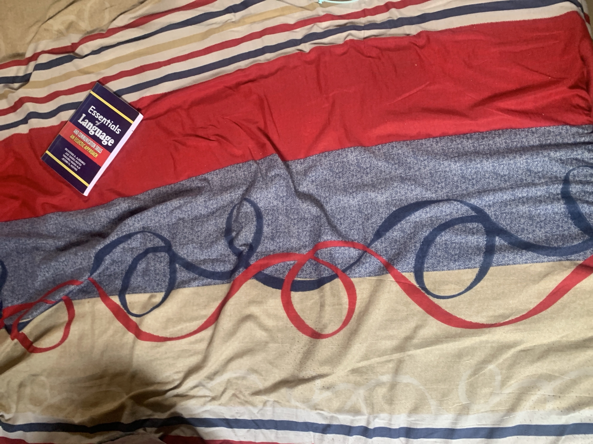 The image shows a book titled "Essentials of Language" placed on a bed with a red, blue, and beige striped pattern. The book is on the red portion of the bed. The bedding is wrinkled. There are no people or specific identifiable landmarks visible. The image does not provide any information about its location in Jauro Yinu, Nigeria.