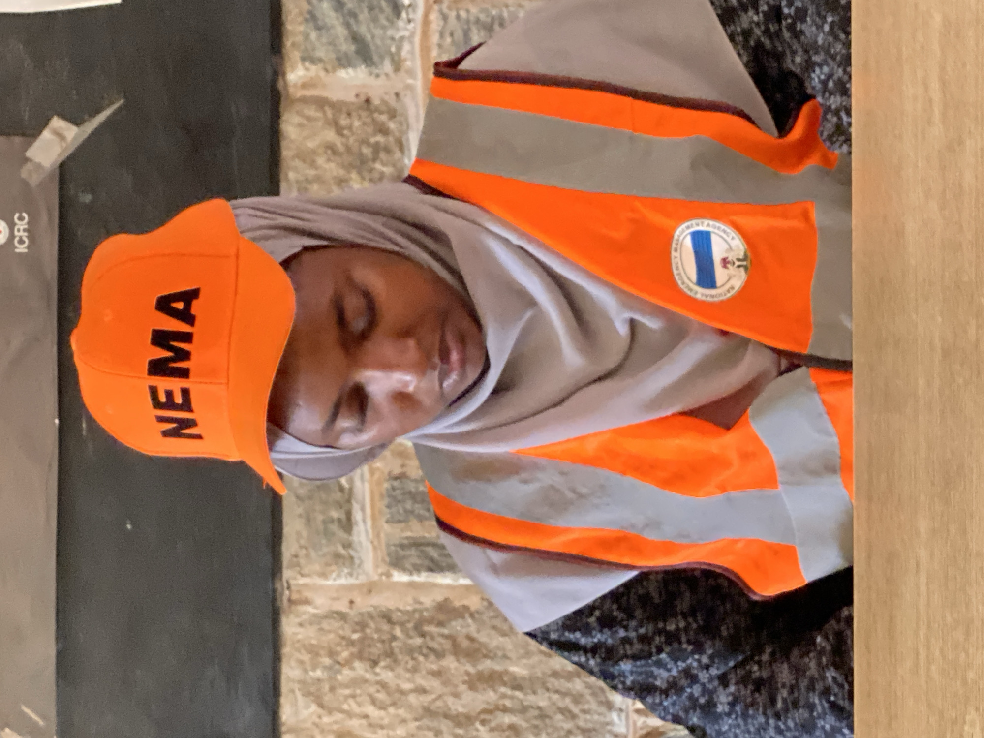 A woman wearing a grey hijab and an orange and grey reflective vest, with a round logo on the right side, and an orange baseball cap with the acronym "NEMA" in black, is oriented diagonally in the frame. The woman appears to be sleeping or resting. To the left of the woman, on a dark grey surface, is a partial view of a document bearing the "ICRC" logo. Behind her, there is a rough stone-like surface, and to the right, a smooth, light brown wooden surface. Based on the logo on her vest, the logo consists of a blue oval in the center depicting a national flag, and circular text around the oval, which confirms that this scene is likely associated with the National Emergency Management Agency (NEMA) in Unguwar Shanu, Nigeria.