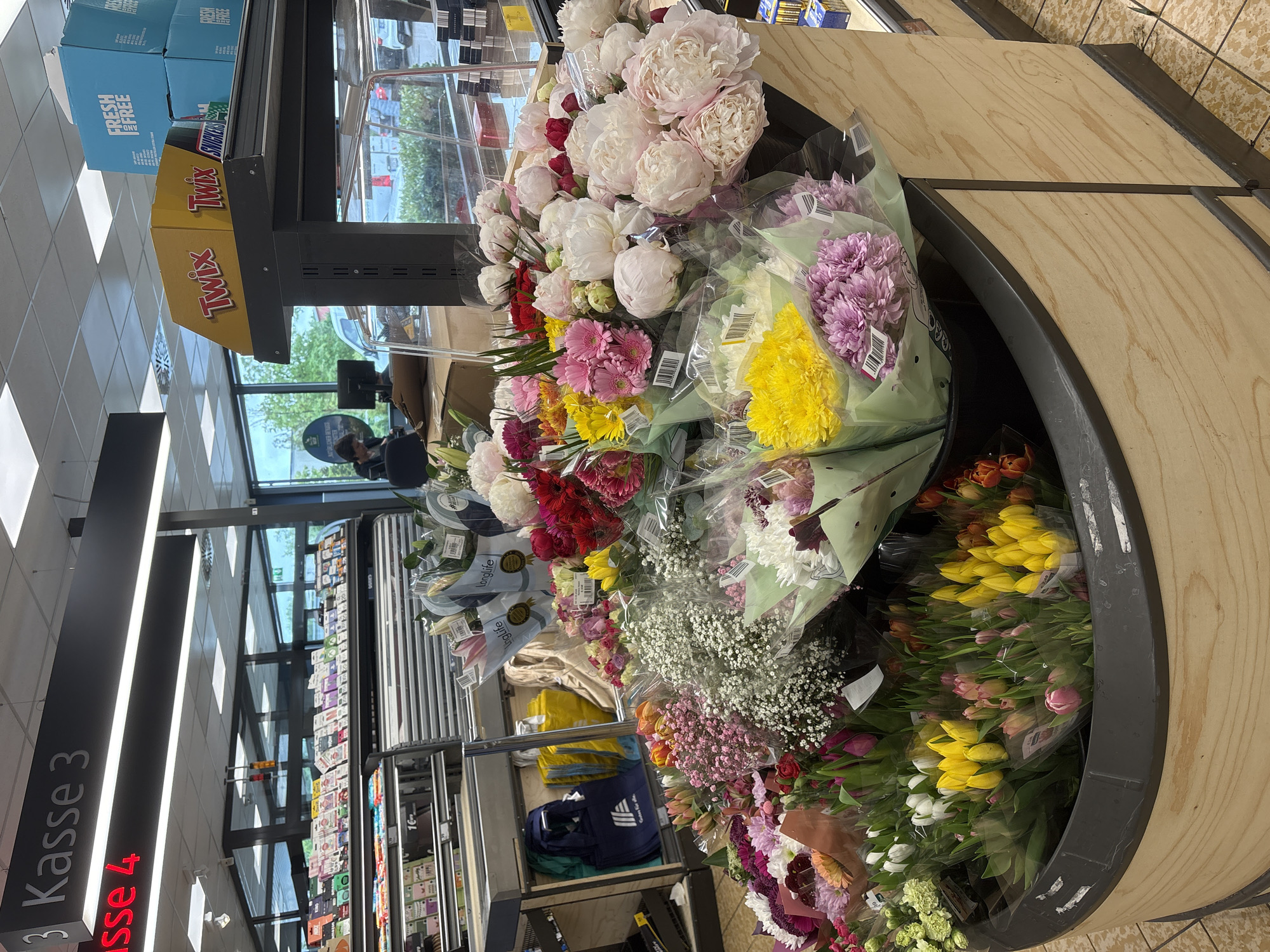 The image depicts a flower stand in a grocery store. The stand is round and contains a variety of flowers including peonies, gerberas, tulips, and baby's breath. There are candy displays for Twix and Snickers. A sign for 'Kasse 3' is visible, along with 'Kasse 4'. The store is located in Kaarst, Germany based on the text 'NATURLICHER GENUSS'.