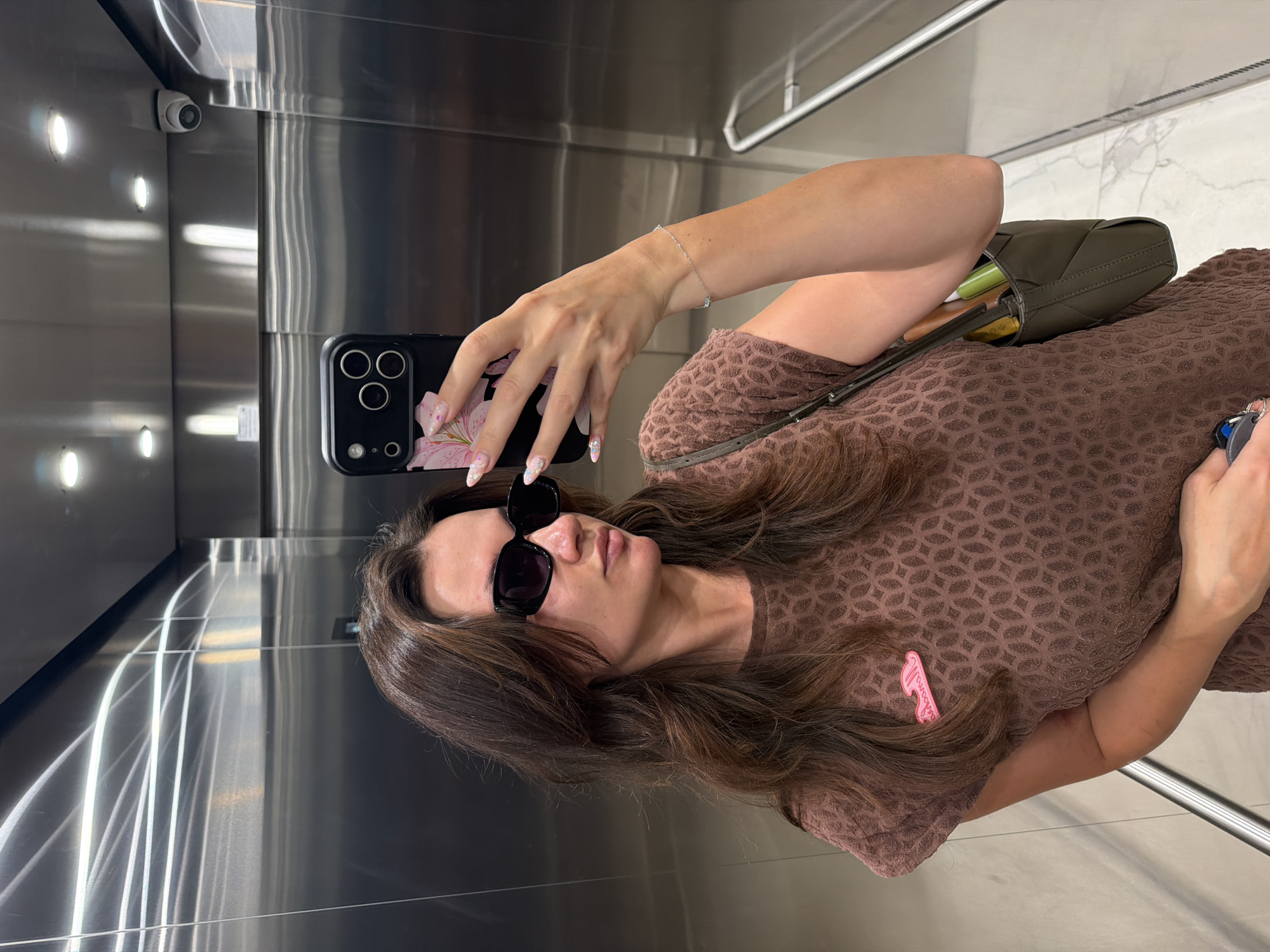 The image shows a woman taking a selfie in what appears to be an elevator. She is wearing sunglasses and a brown patterned top, and is holding a phone in her right hand. A green purse is slung over her shoulder. A security camera is visible on the wall to her left, and the elevator walls are made of brushed metal. The lighting suggests it is daytime. A pink hairpin is attached to her shirt.