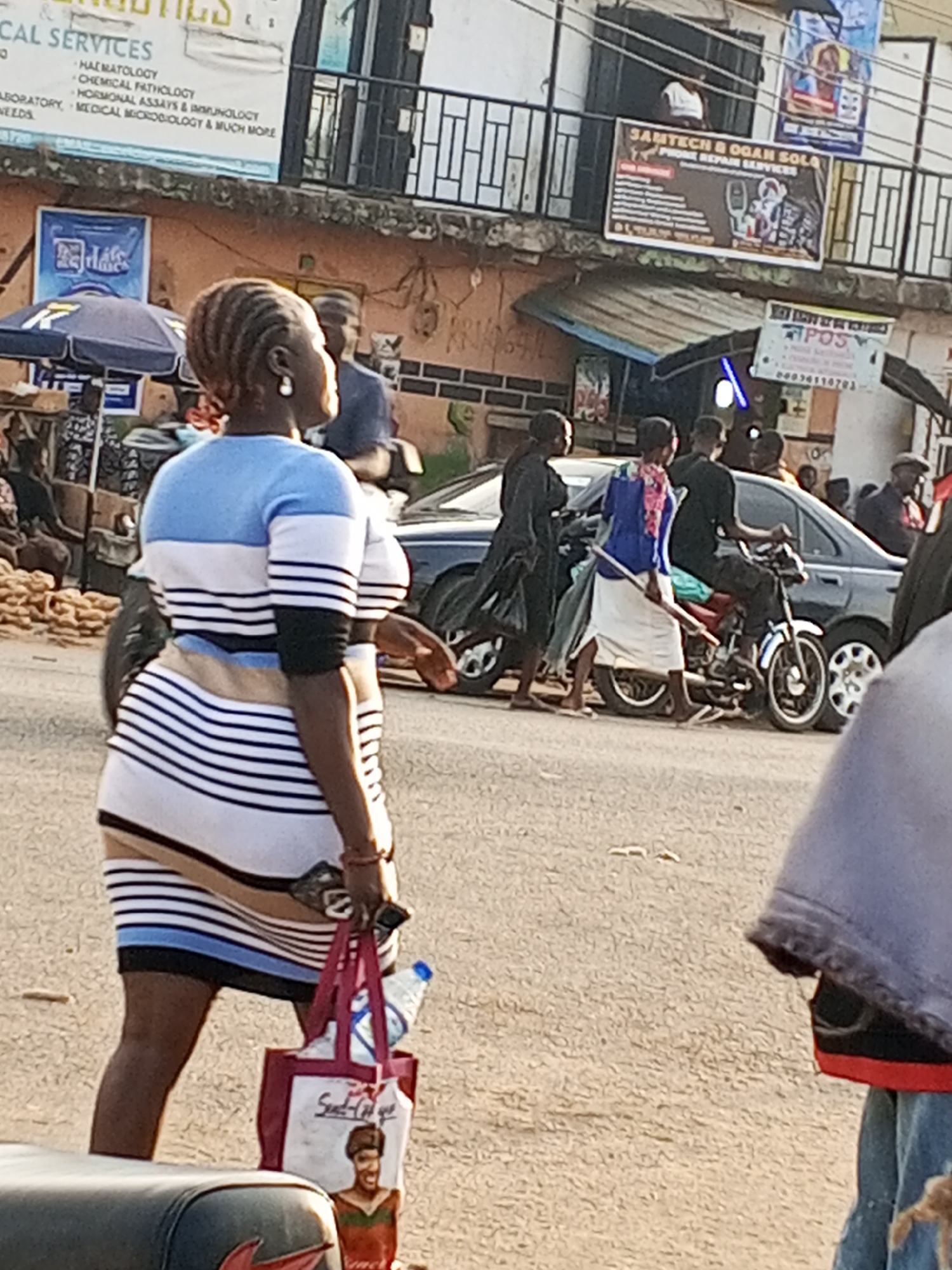 The scene is on a street in Gwong, Nigeria. An African woman with short, braided hair and wearing a blue, white, and black striped dress is walking towards the camera, carrying a red and white shopping bag. Behind her, a black sedan is parked on the side of the road. A man is seated on a parked red motorcycle next to the car. Several people are walking on the sidewalk. In the background, there are buildings with advertisements on the upper levels. These include a sign for medical services (HAEMATOLOGY, CHEMICAL PATHOLOGY, etc.) and a phone repair service called "SAMTECH & OGAN SOLQ". A "POS" sign is also visible. The sky is overcast.