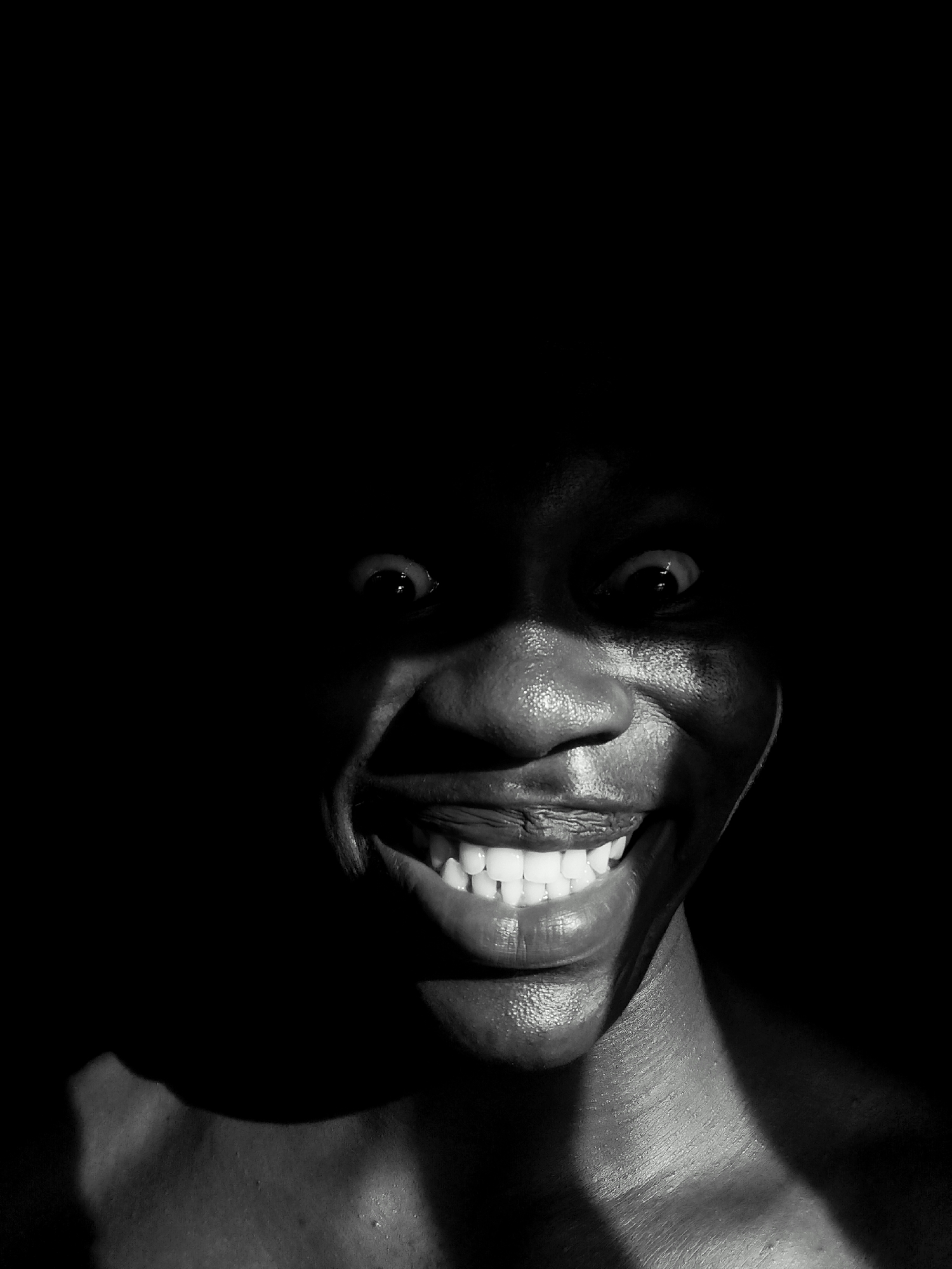 A black and white, close-up portrait captures a person with dark skin in stark lighting. The person is smiling widely, revealing white teeth, and their eyes are wide. A dark area surrounds the person's head. The lighting creates strong contrasts, highlighting the person's facial features and parts of their upper body. The location is not discernable from this image, but is specified as Keffi, Nigeria.