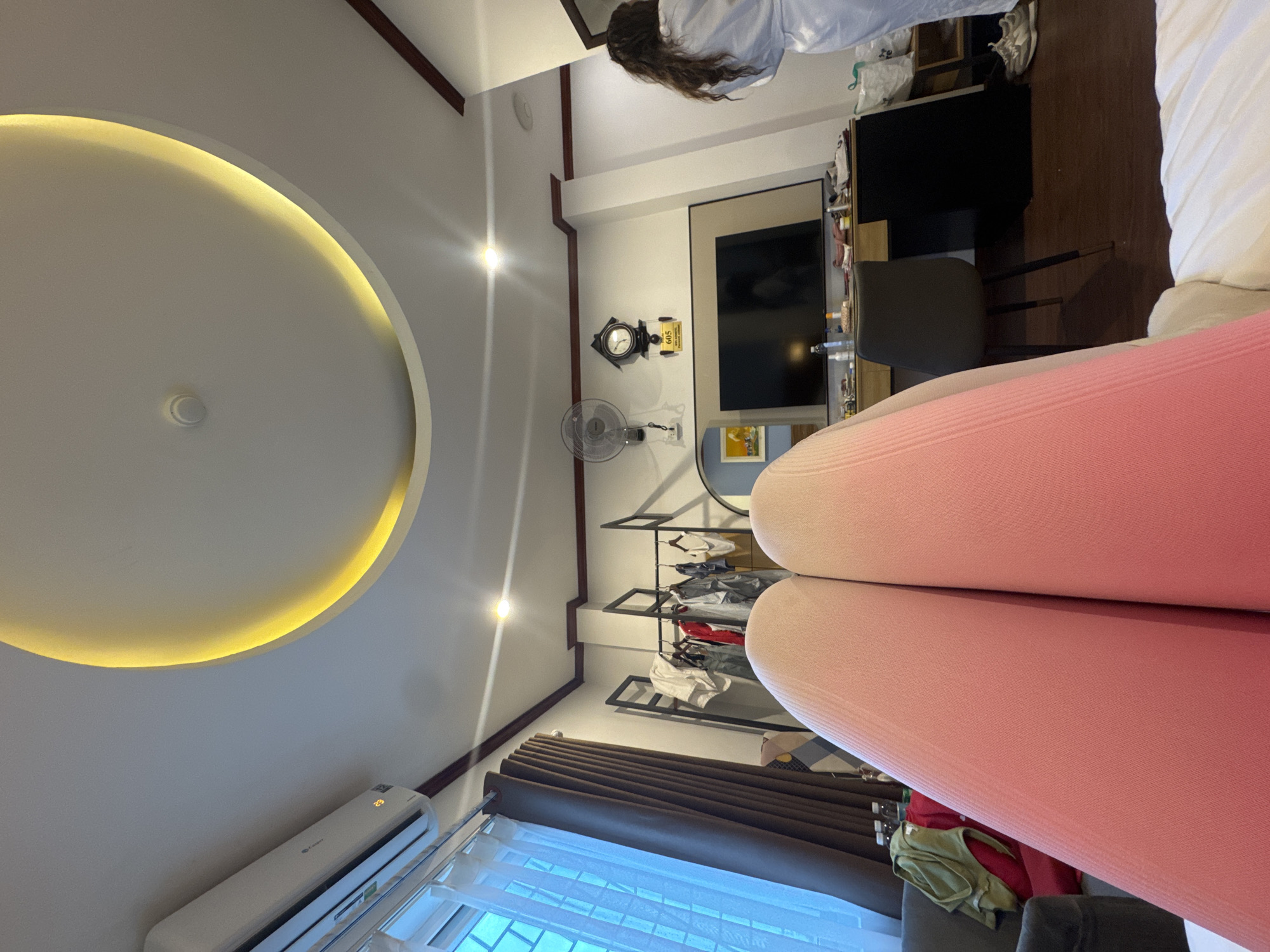 The image depicts an indoor scene, possibly a hotel room, in Hà Nội, Vietnam. In the foreground, two legs wearing pink leggings are visible, with a bed in the lower right corner. The room features an air conditioning unit mounted on the ceiling, a circular illuminated ceiling design, a wardrobe rack with clothes, a desk with a television, a chair, and a wall clock. A person wearing a white robe is standing on the right side of the frame, with their back to the camera. The overall impression is a casual, lived-in space.