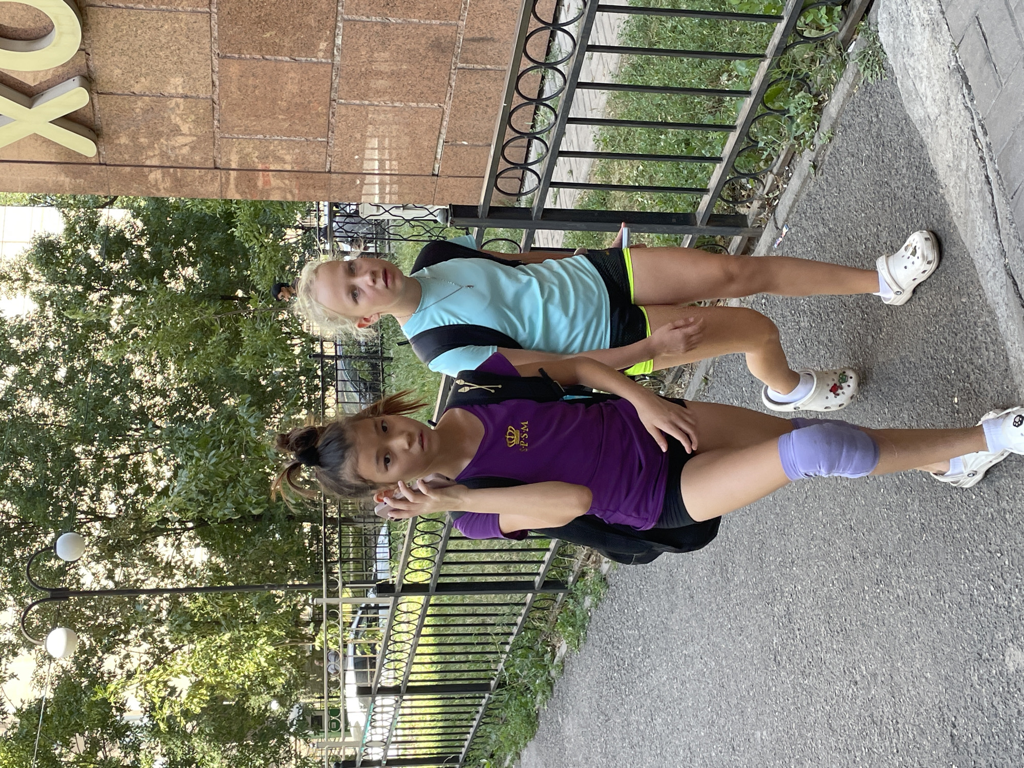 Two young girls are walking on a sidewalk next to a metal fence and a building with a large sign. The girl on the left has her hair pulled up in a bun and is wearing a purple t-shirt and black shorts, with a knee pad on her left leg. She is holding a phone to her ear. The girl on the right has blonde hair and is wearing a light blue t-shirt and black shorts with yellow trim. She is carrying a black backpack. The building behind them has a large, yellow sign with Cyrillic lettering. The environment appears to be urban, with trees and greenery visible. The sign on the building indicates this location is likely in Almaty, Kazakhstan, specifically in front of a building that has the sign "XO" which is visible on the building.