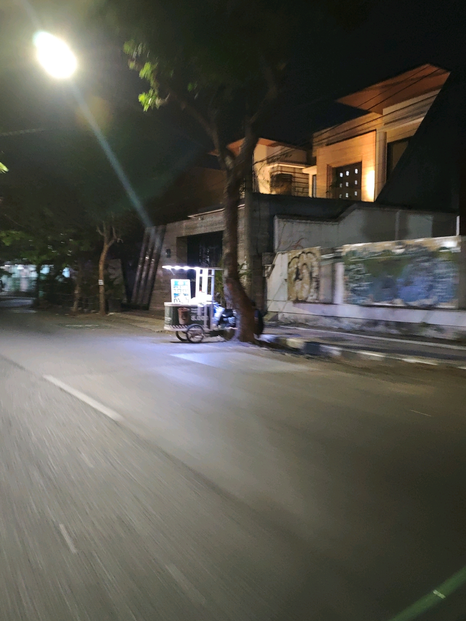 The image shows a street scene at night in Kediri City, Indonesia. A food cart with a lit display is parked on the side of the road, illuminated by streetlights. Behind the cart is a building with modern architecture, featuring large windows and a distinctive roofline. The street itself is asphalt, with a white line marking the edge. Trees line the sidewalk. The image is slightly blurred, suggesting motion, possibly from a vehicle.