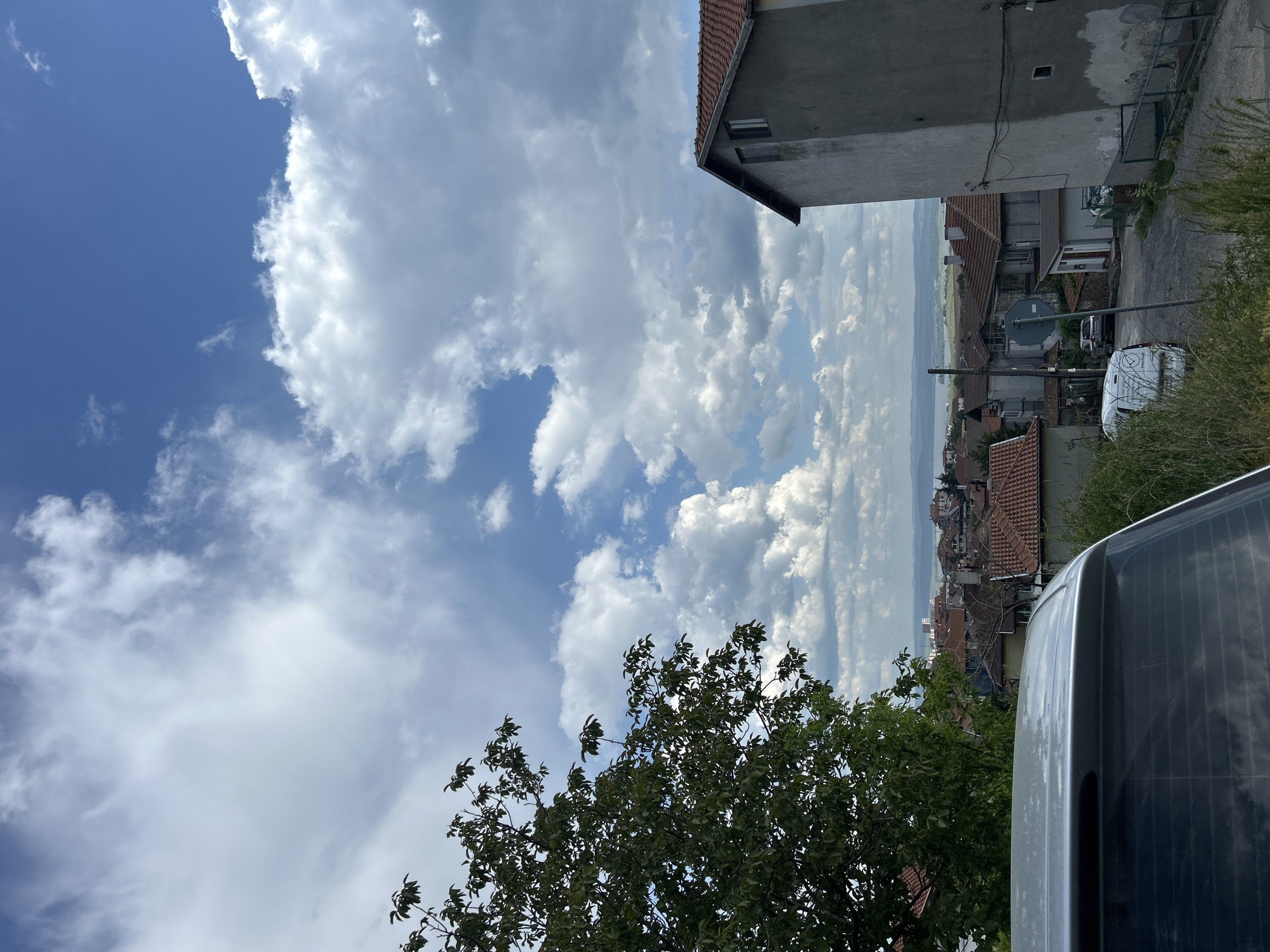 The image shows a view in Burgas, Bulgaria. The foreground includes the top of a car, a tree, and part of a building. Beyond that, there's a view down a street towards a skyline with many buildings and tile roofs, stretching toward a distant horizon line. The sky is partly cloudy.