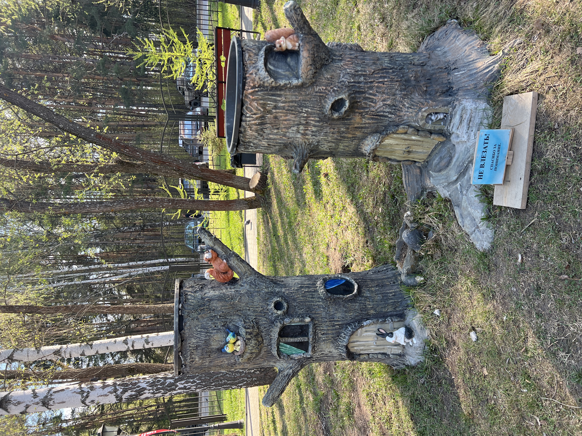The image shows two decorative tree stump structures in a park in the city of Chaikovsky, Russia. The tree stumps appear to be made of artificial material and are decorated with animal figurines such as squirrels, rabbits, and birds. One stump has a trash receptacle in the top. A sign near one of the stumps reads "Do not climb! Thank you for your understanding." The background features trees, grass, and a fence.