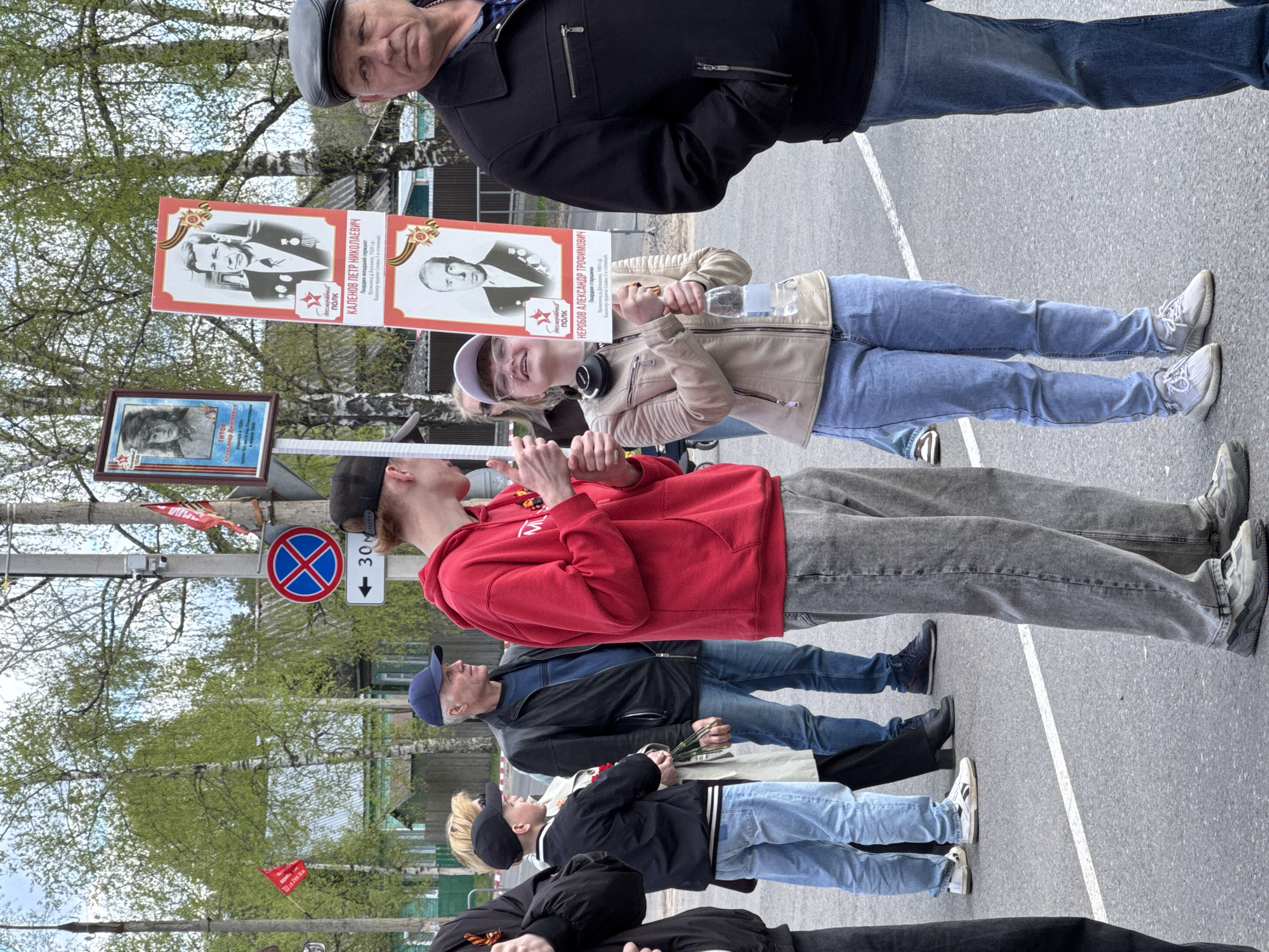 In Yamyshevo, Russia, a group of people are participating in a march, likely a memorial event. They are holding portraits of military personnel. The participants are dressed in casual clothing, including jeans, jackets, and hoodies. A road sign is visible in the background, indicating traffic restrictions. The environment suggests an urban area with trees and buildings in the periphery.