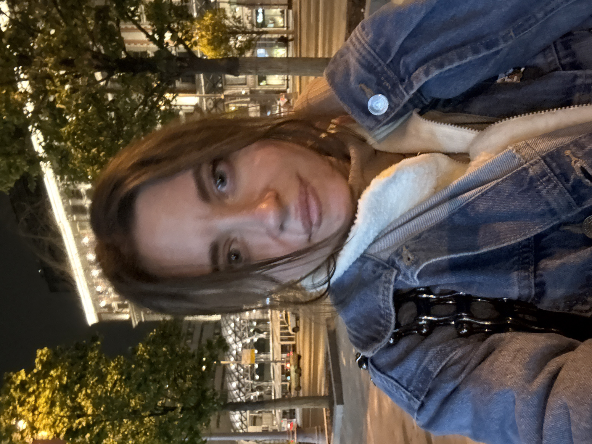 A selfie of a woman wearing a blue denim jacket with a white, furry lining. The background shows a cityscape at night, with trees on the left and buildings illuminated with lights. The location is Moscow, Russia, identifiable by the GUM department store in the distance.