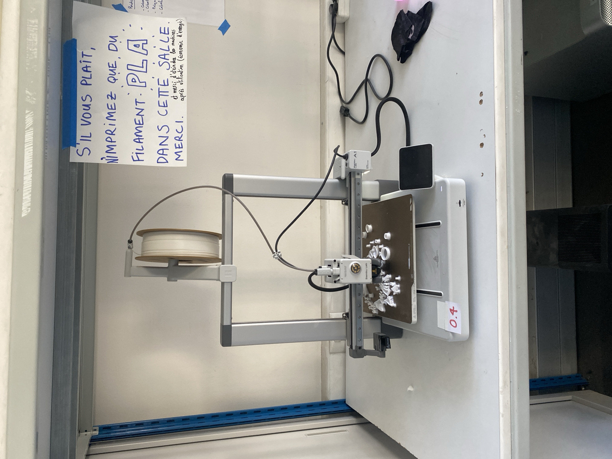 The image displays a 3D printing station, likely situated in a workshop or shared lab in Paris, France. A Bambu Lab A1 Mini 3D printer, identifiable by the "Bambu Lab" and "A1" markings, is positioned prominently. A spool of white or light-colored filament is mounted on the printer's holder, and its print bed is covered with several small, white 3D-printed objects, indicating recent or ongoing use. A red sticker with "0.4" is attached to the print bed, probably signifying the nozzle size.
A handwritten note in French is taped to the wall above the printer. It instructs users: "S'IL VOUS PLAÎT, N'IMPRIMEZ QUE DU FILAMENT: PLA DANS CETTE SALLE MERCI." (Please, only print with PLA filament in this room. Thank you.) The note further advises: "et merci d'éteindre les machines après utilisation (économie d'énergie)" (and thank you for turning off the machines after use (energy saving)). A partial, less legible list of words like "Poids," "Color," and "Poly" is visible at the top right of the note.
The printer is connected to power outlets via black cables and cords. A black object, possibly a glove or cloth, is placed near the cables. Below the printer, a blue metal slotted rail is affixed to the wall. The surroundings are brightly lit, suggesting an indoor environment, possibly a clean and functional educational or communal maker space.