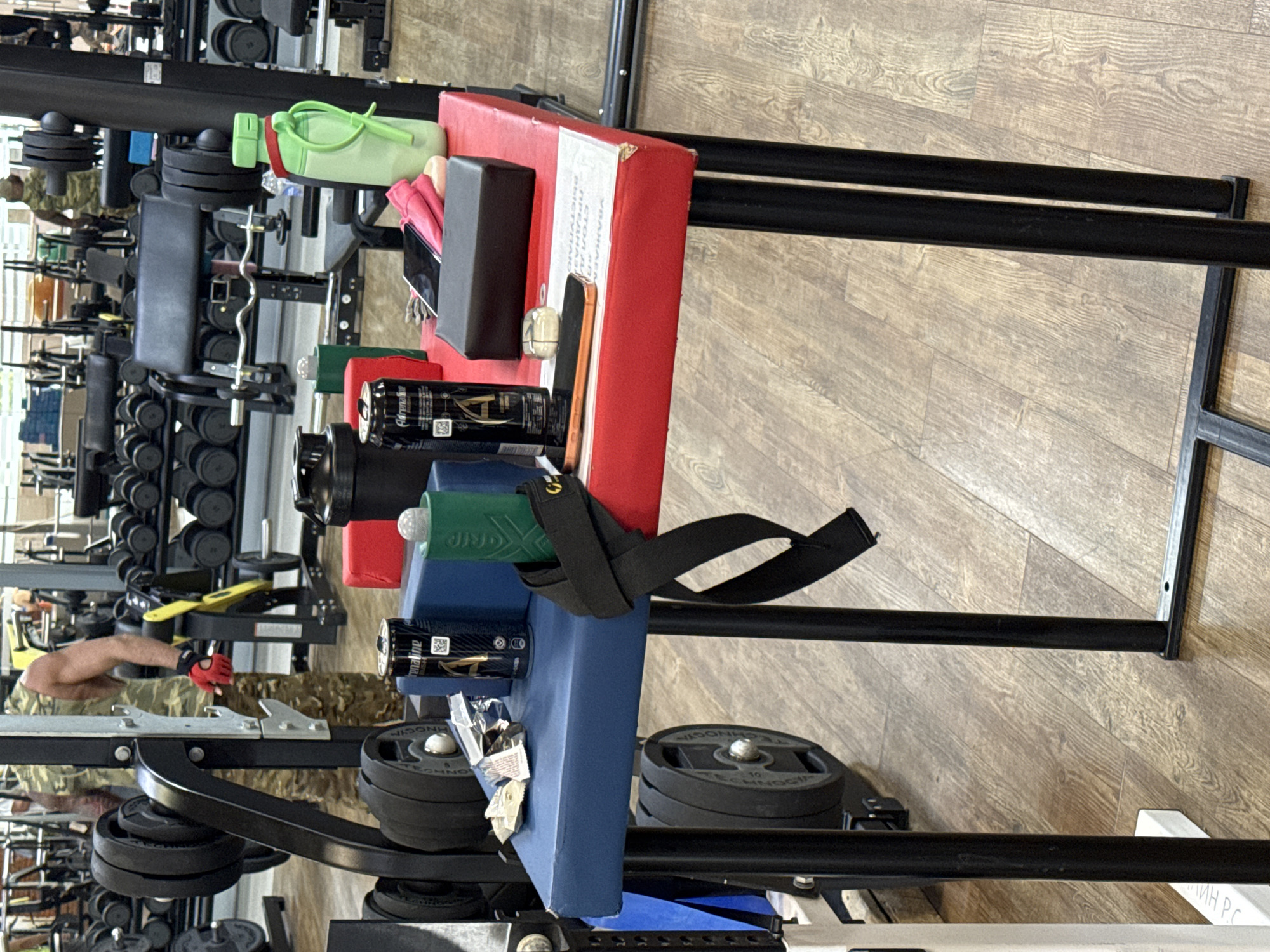 The image shows an arm wrestling table set up in a gym. The table has a red top and a blue padded surface for the arms. On the table are several objects, including a can of "Adrenaline Rush" energy drink, a green grip trainer, a light green water bottle, a black smartphone, a pair of pink earbud cases, a black rectangular object, and some wrappers from a snack. A black arm wrestling strap is visible on the side of the table. In the background, there are weightlifting machines and dumbbells, and a person wearing camouflage clothing is visible. The floor is made of wooden planks. The location is identified as a gym in the city of Chaikovsky, Russia, suggesting the gym is likely a local facility within the city. The scene is static, with no immediate action taking place.