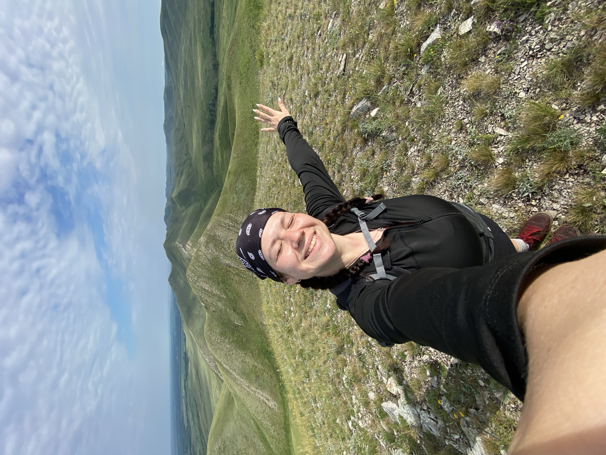 A smiling woman with braided hair, wearing a black bandana and black jacket, takes a selfie on a grassy, rocky hillside with a panoramic view of rolling green hills under a partly cloudy sky. She is extending her right arm towards the camera, with her palm open. The landscape appears to be in a rural or mountainous region, possibly the area around Verkhnyaya Chernorechka, Russia, given the topography.