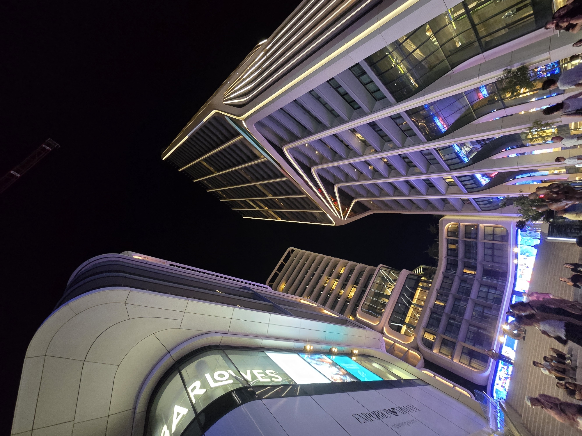 An upward-angle night shot captures the contemporary architecture of a modern complex in Saint Julian's, Malta. The buildings are characterized by sleek, curved lines and are illuminated with bright, white and purple lights that highlight their structural features and balconies. The facade of one building features a large digital screen displaying what appears to be advertisements or artistic visuals, with the text "AR LOVES" prominently displayed. Another building has the logo "EMPORIO ARMANI" visible on its exterior. In the foreground, a wide staircase leads up to the complex, with several individuals seated or standing on the steps. The overall atmosphere is lively, suggesting a public space or shopping district at night. The sky is dark, indicating nighttime.